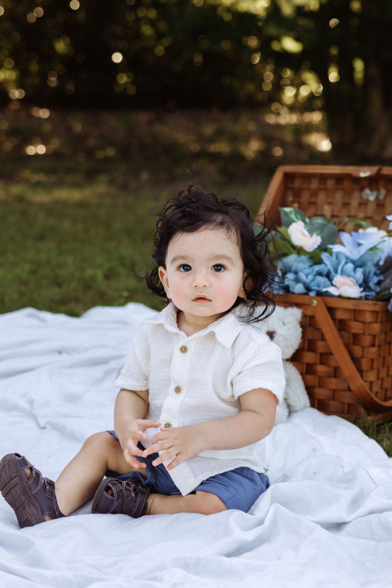 Baby in white outfit sitting on blanket with wicker basket and blue flowers outdoors.