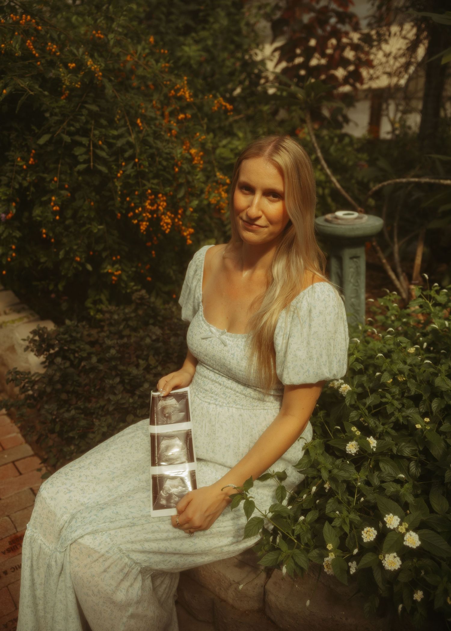 Pregnant woman in floral dress holding ultrasound photos while sitting in a garden surrounded by greenery.