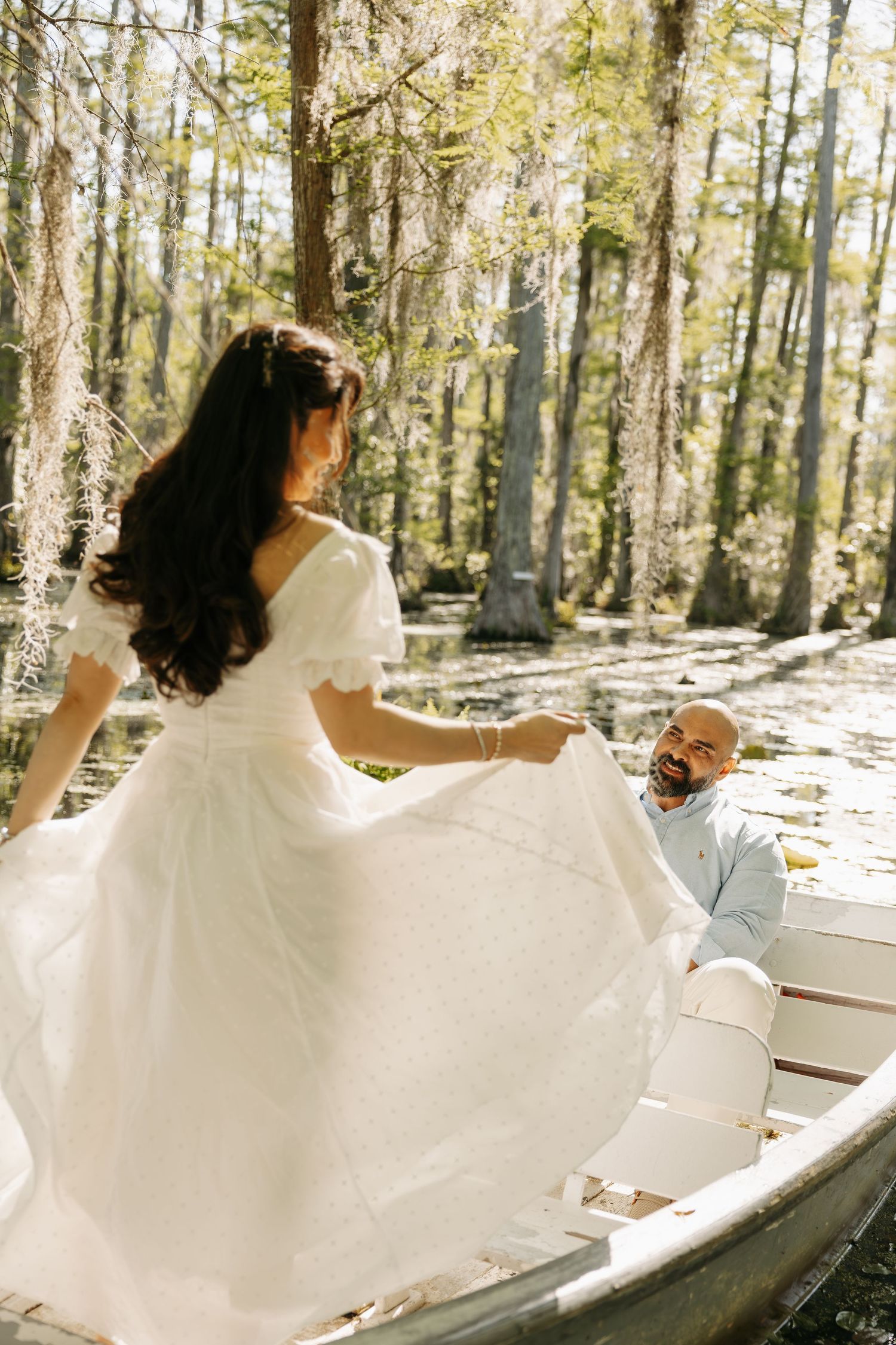 Bride in white off-shoulder gown twirling on a boat in a sunlit cypress swamp, groom smiling up at her.