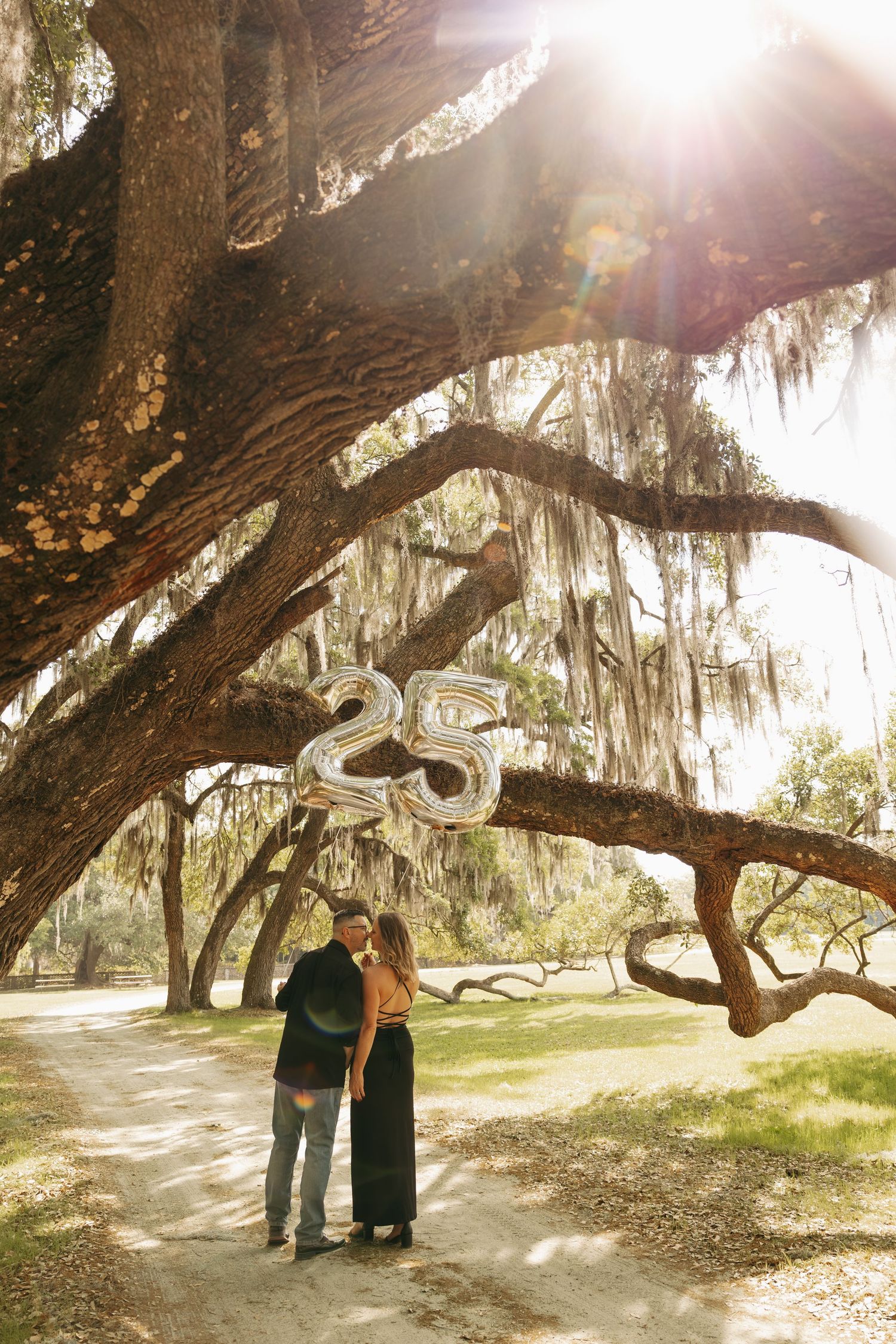 Couple embracing under sprawling oak trees draped in Spanish moss, holding silver '25' balloons in a sunlit park.