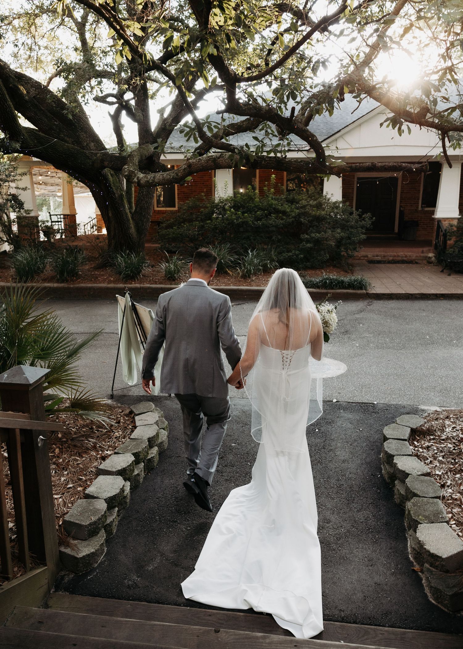 Bride and groom walk hand-in-hand toward a charming rustic venue surrounded by trees at golden hour.