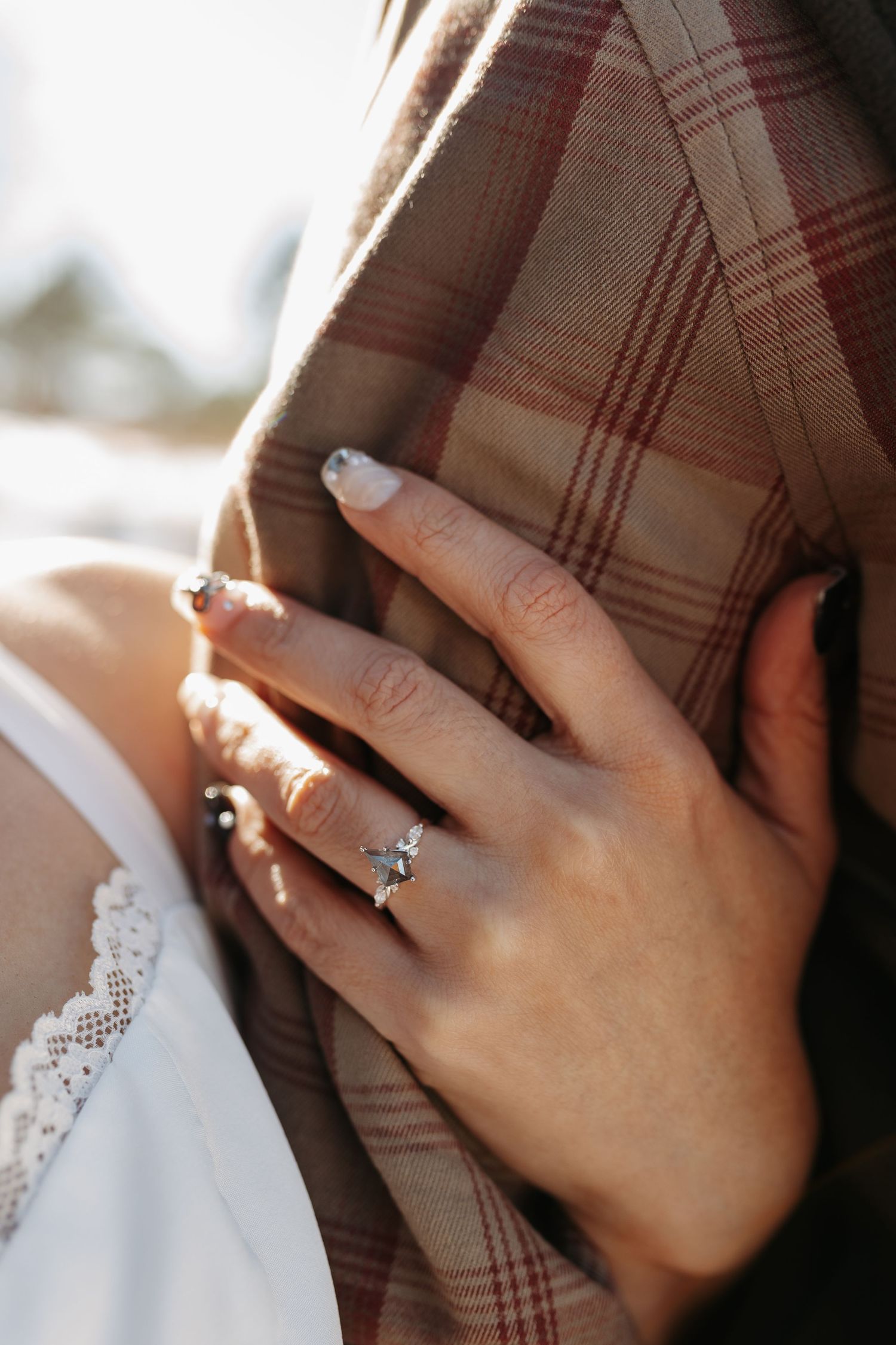 Close-up of a woman's hand with elegant nails and a diamond engagement ring resting on a plaid jacket.