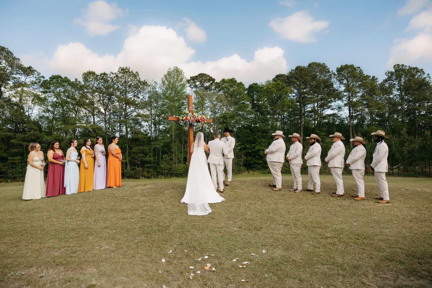 Wedding ceremony outdoors with bridesmaids in colorful dresses and groomsmen in tan suits by wooden cross.