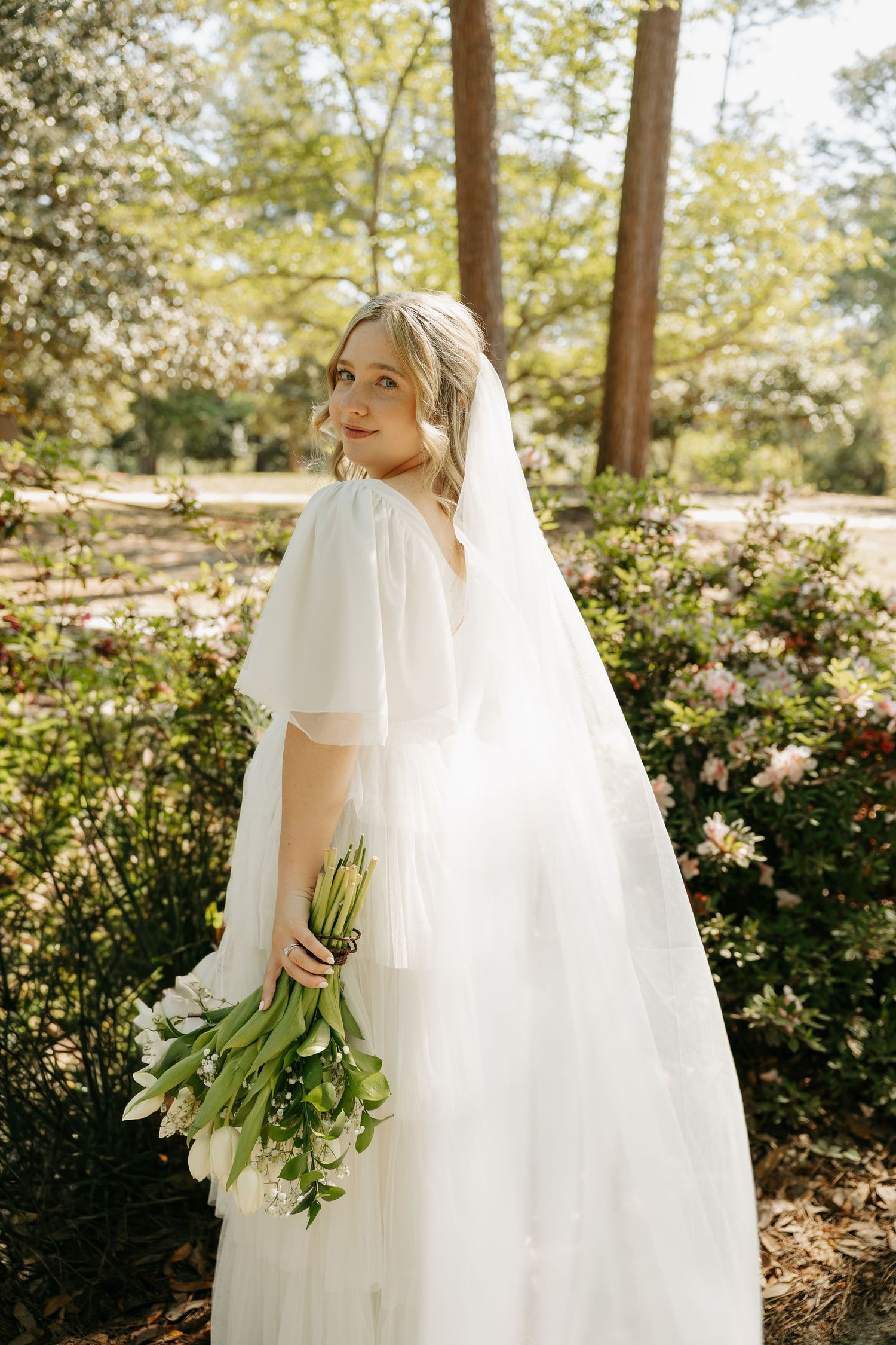 Bride in white flutter-sleeve gown and veil holds white tulip bouquet, posing in a lush garden setting.