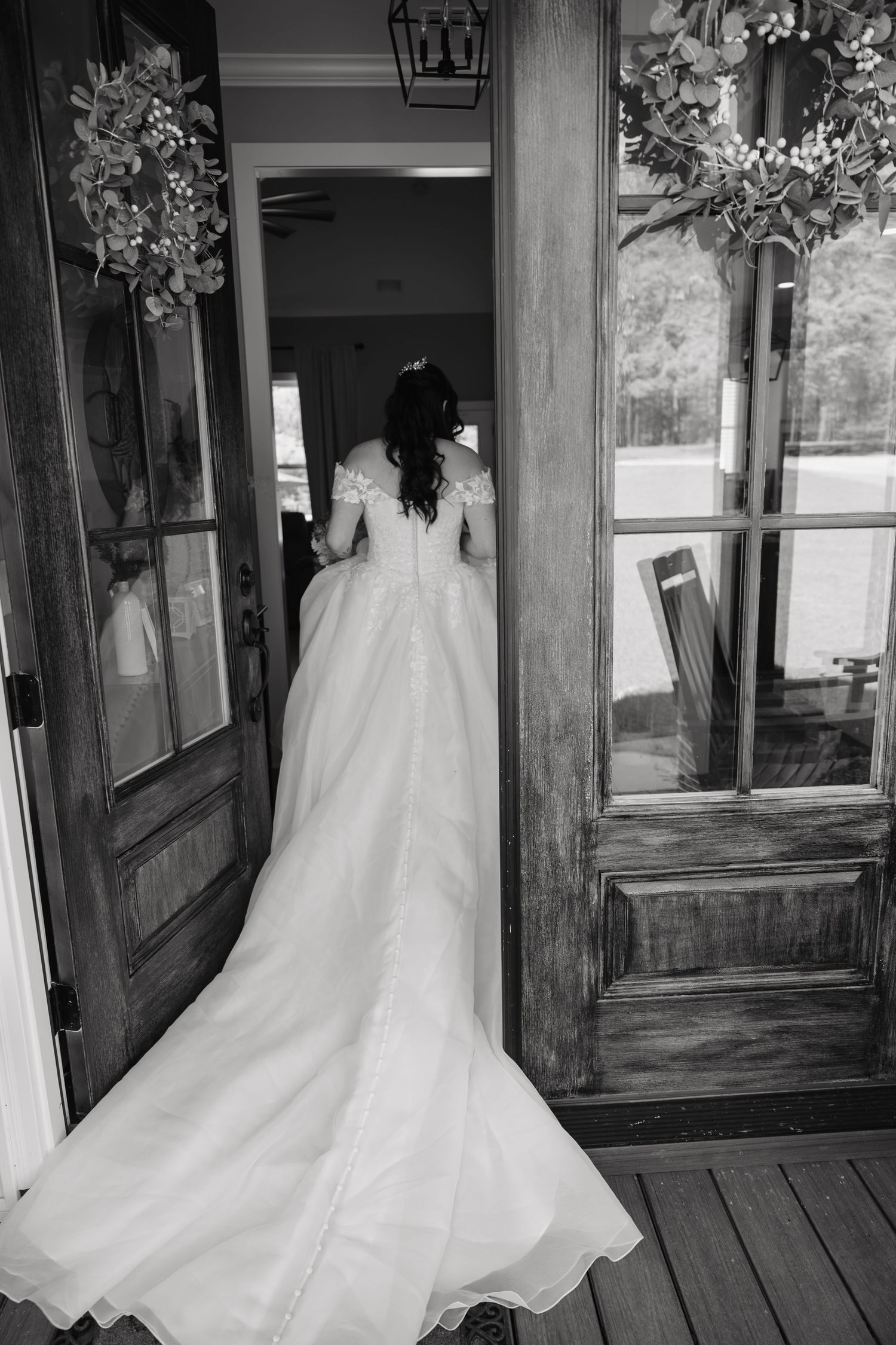 Black and white photo of a bride in a flowing wedding gown standing in rustic double doors adorned with floral wreaths.