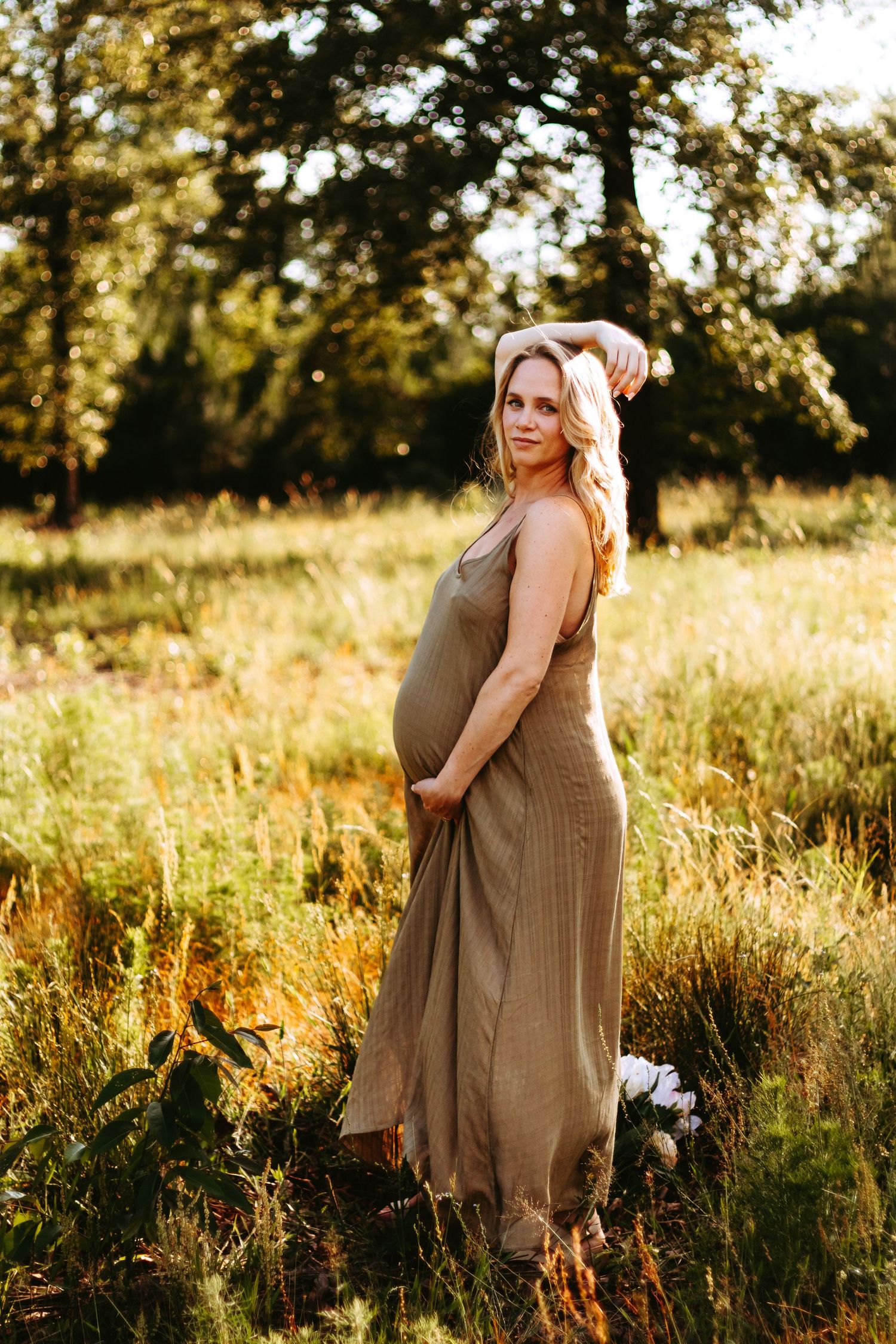 A maternity portrait in a brown dress set against a golden sunset in a summer meadow.