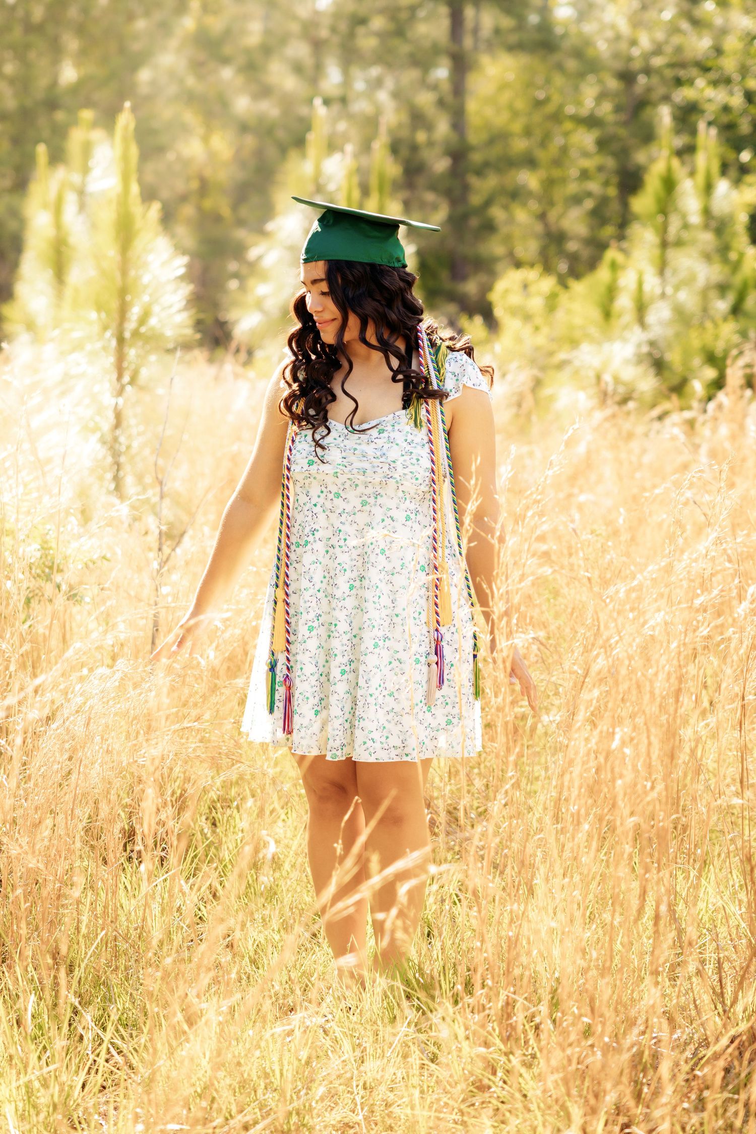 Graduate in floral dress and cap walks through tall golden grass at sunset.