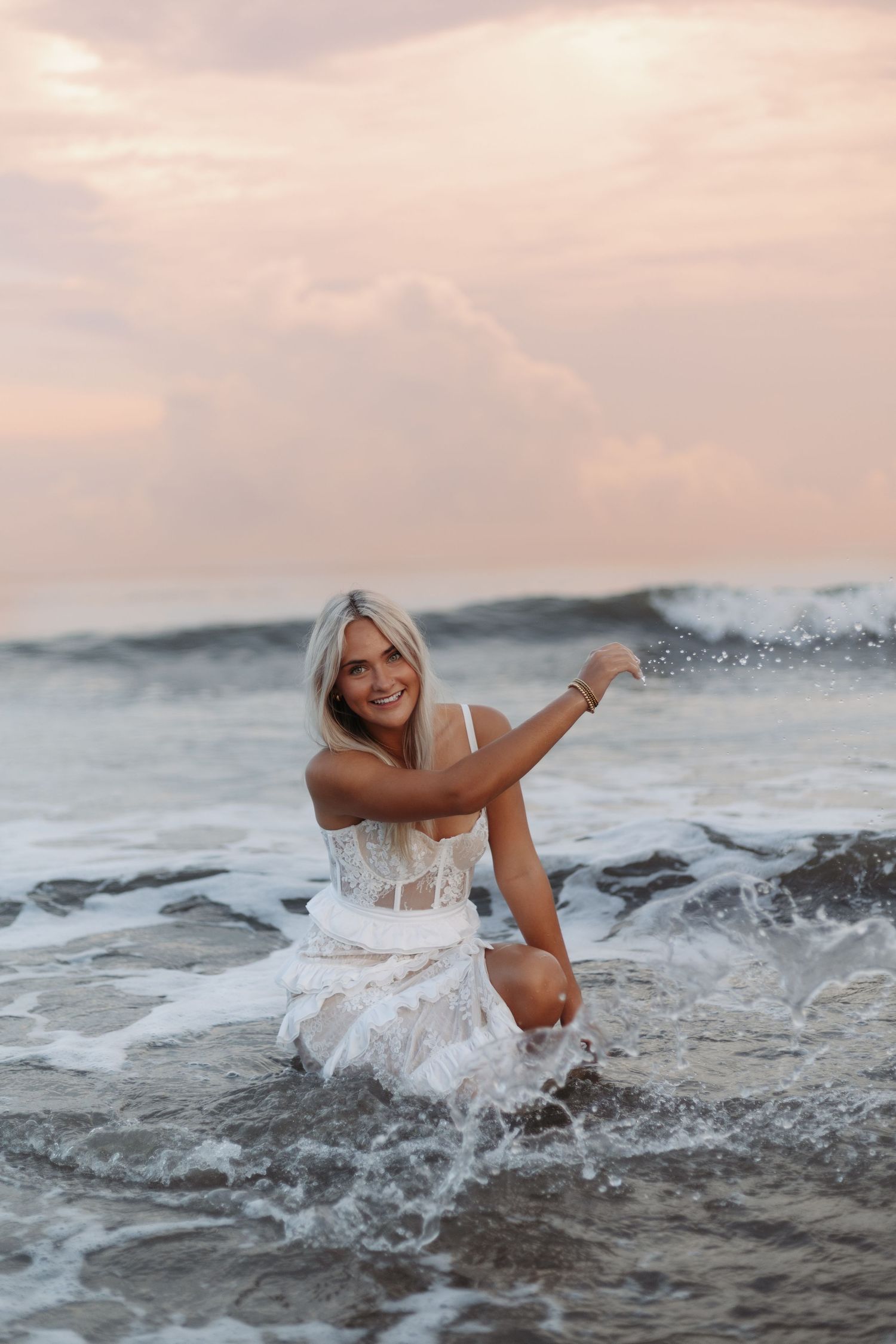 A person in a white lace dress sits on coastal rocks at sunset as ocean waves splash around them.