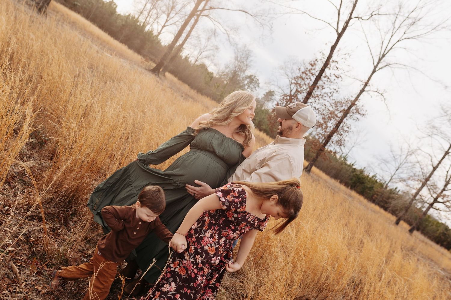 Family of four posing in a golden field with bare trees, wearing boho-style outfits including floral and olive green dresses.