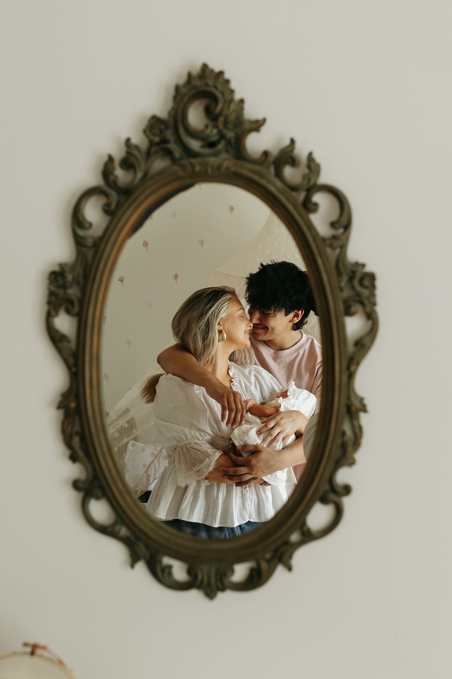 Couple embracing and kissing reflected in an ornate vintage oval mirror mounted on a white wall.