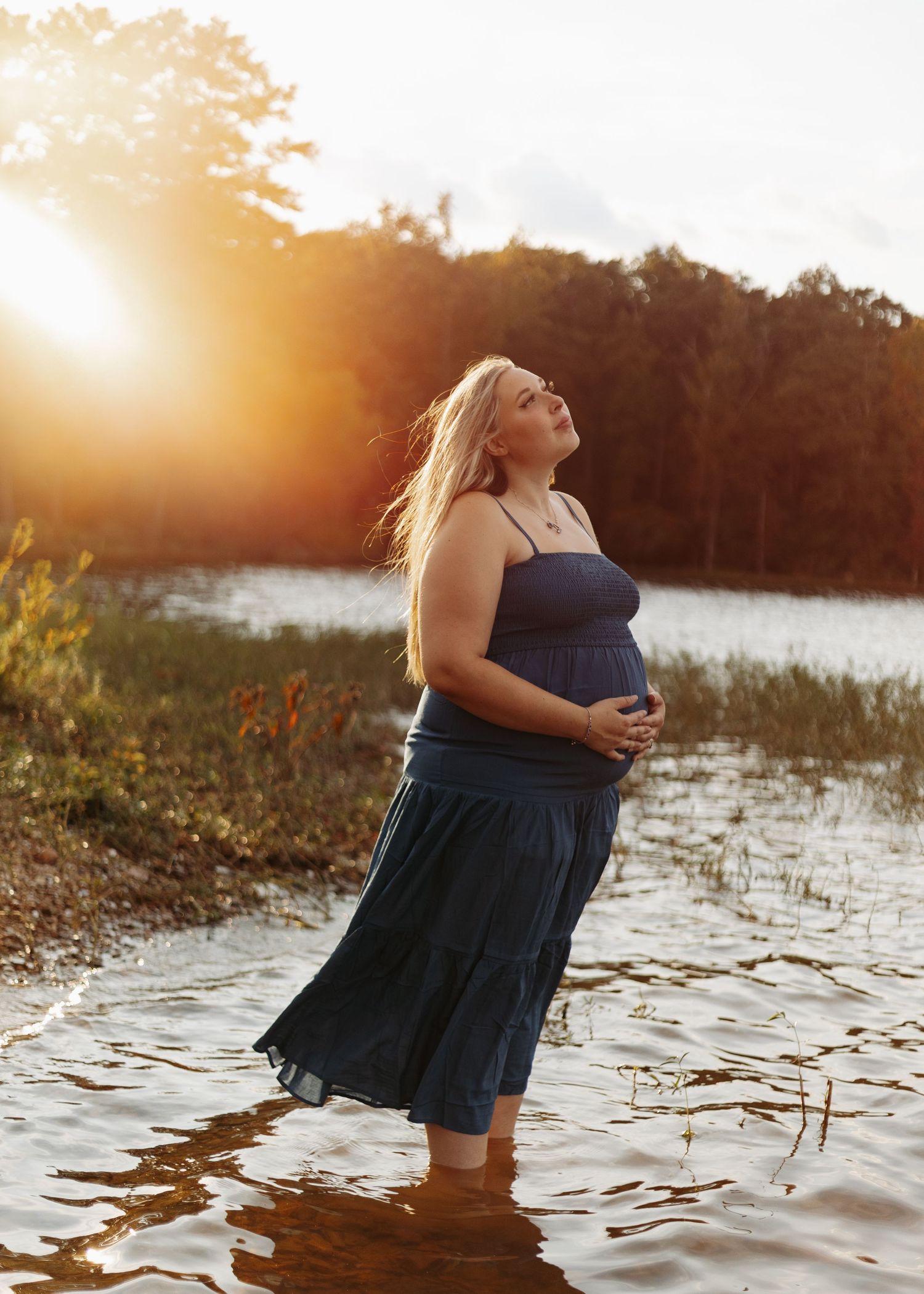 Figure in a blue dress stands at the shoreline during golden hour with sun flare and reflective water.