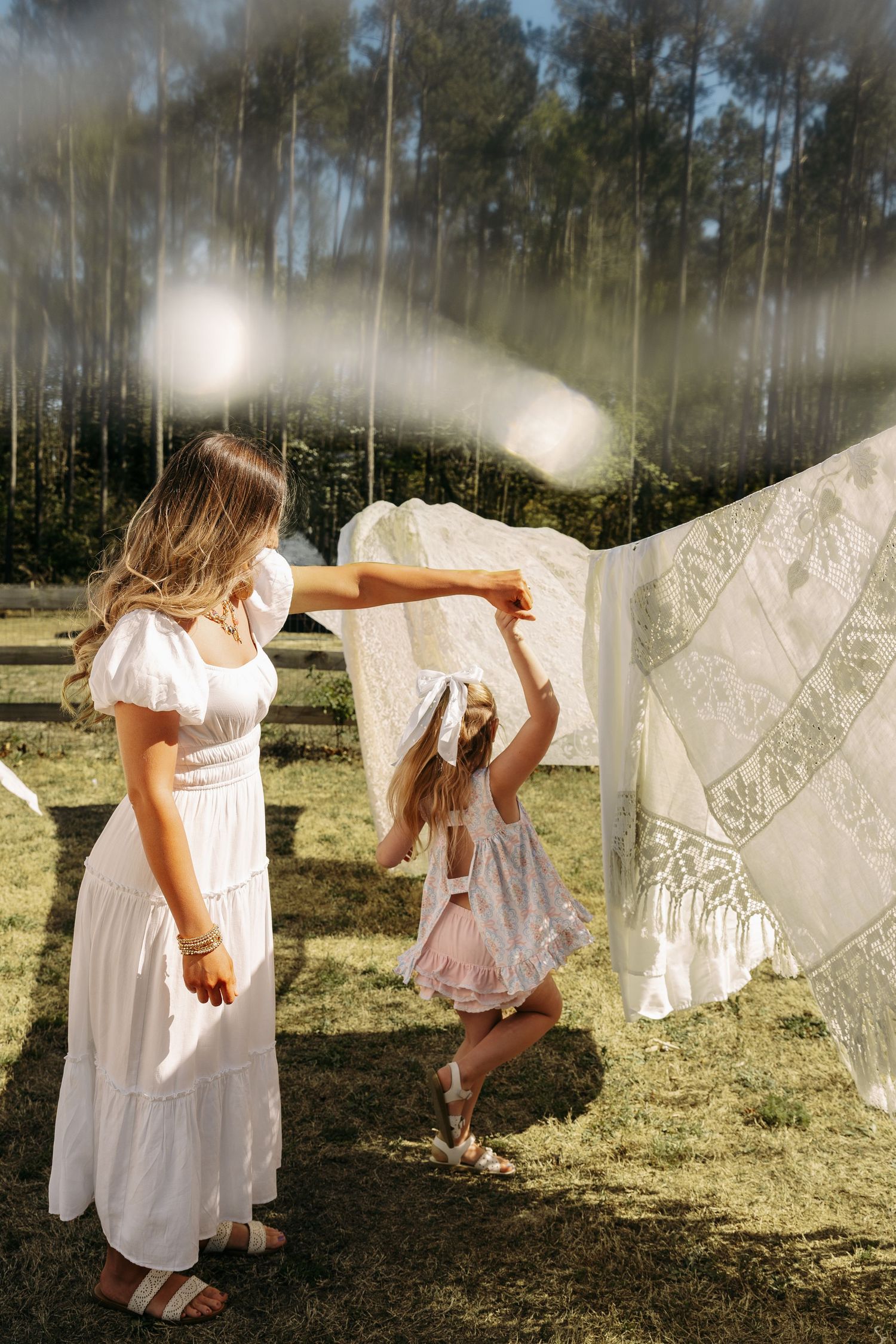 A woman and young girl in white dresses play with hanging laundry in a sunlit outdoor field surrounded by trees.