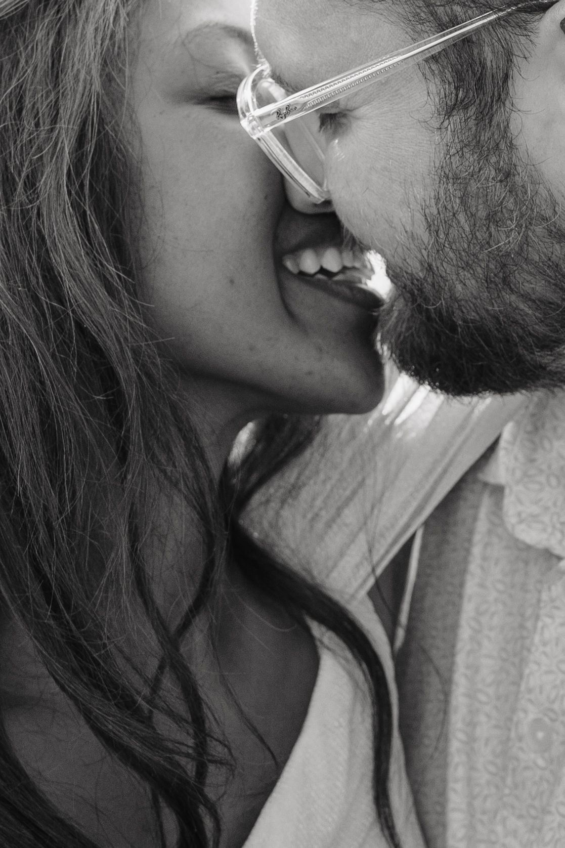 Black and white photo of a smiling couple sharing a romantic kiss, man wearing glasses with a beard.