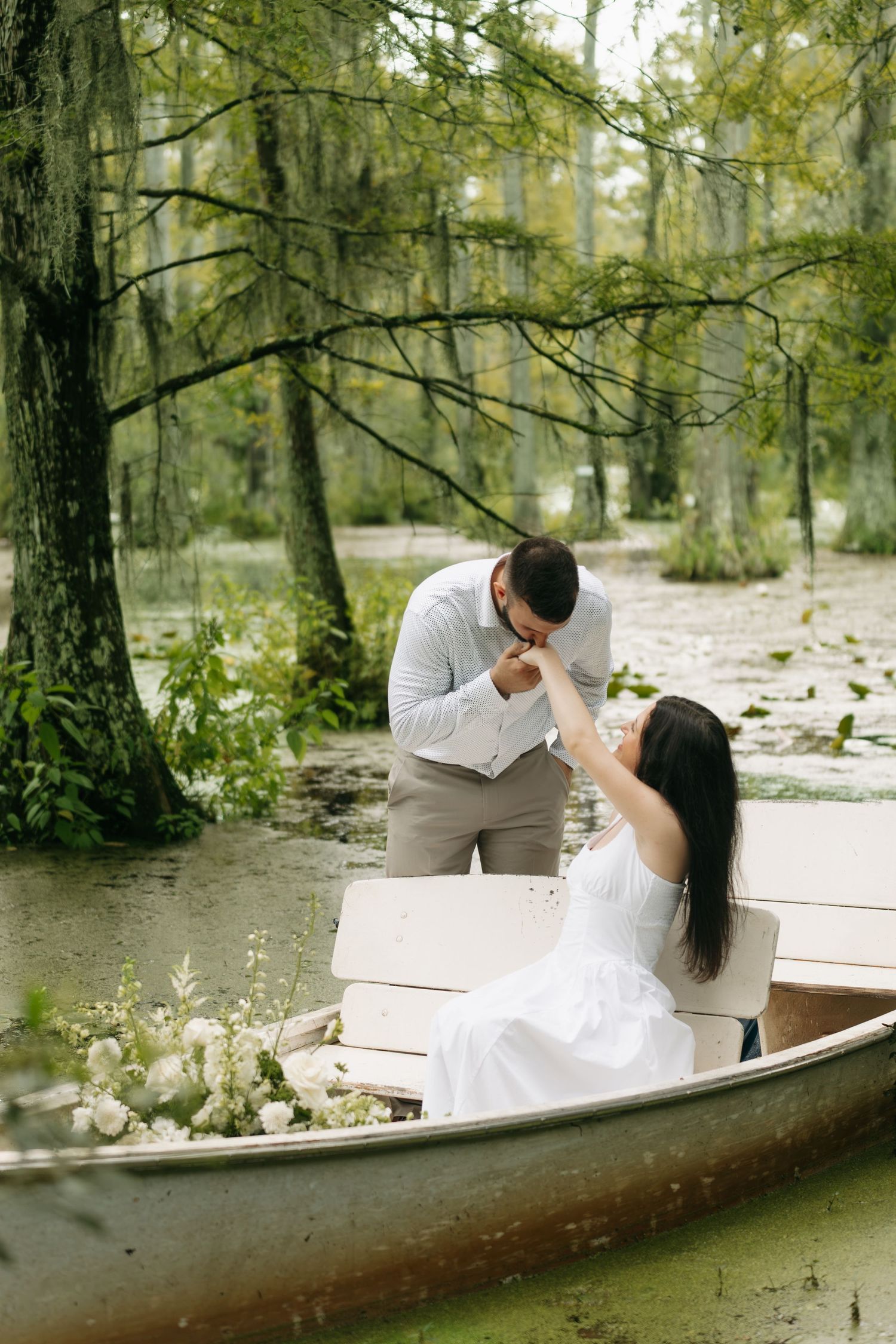 A groom kisses his bride's hand while she sits in a white boat surrounded by lush greenery and white flowers.