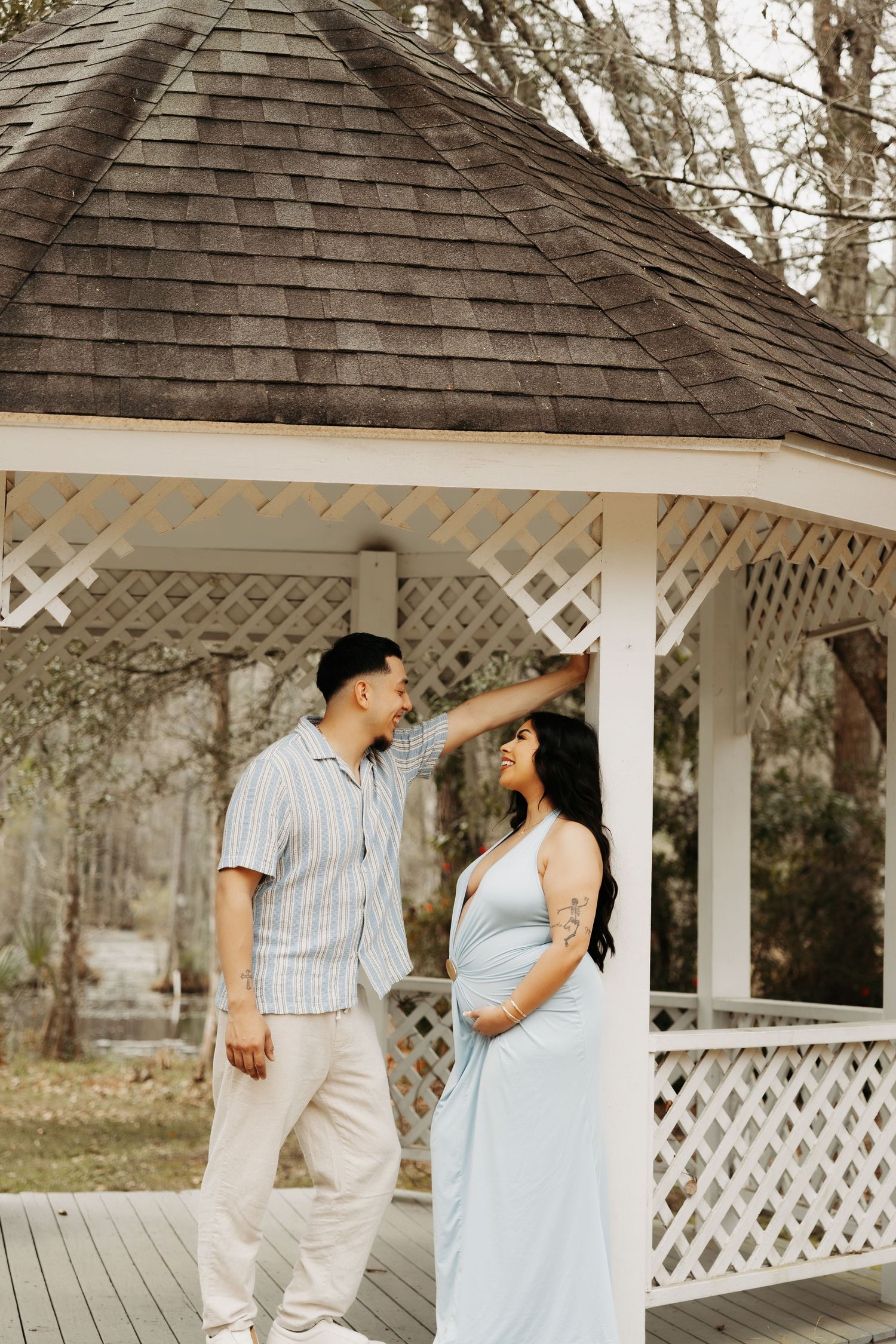 Expecting couple smiles together by a white gazebo in a serene outdoor setting.
