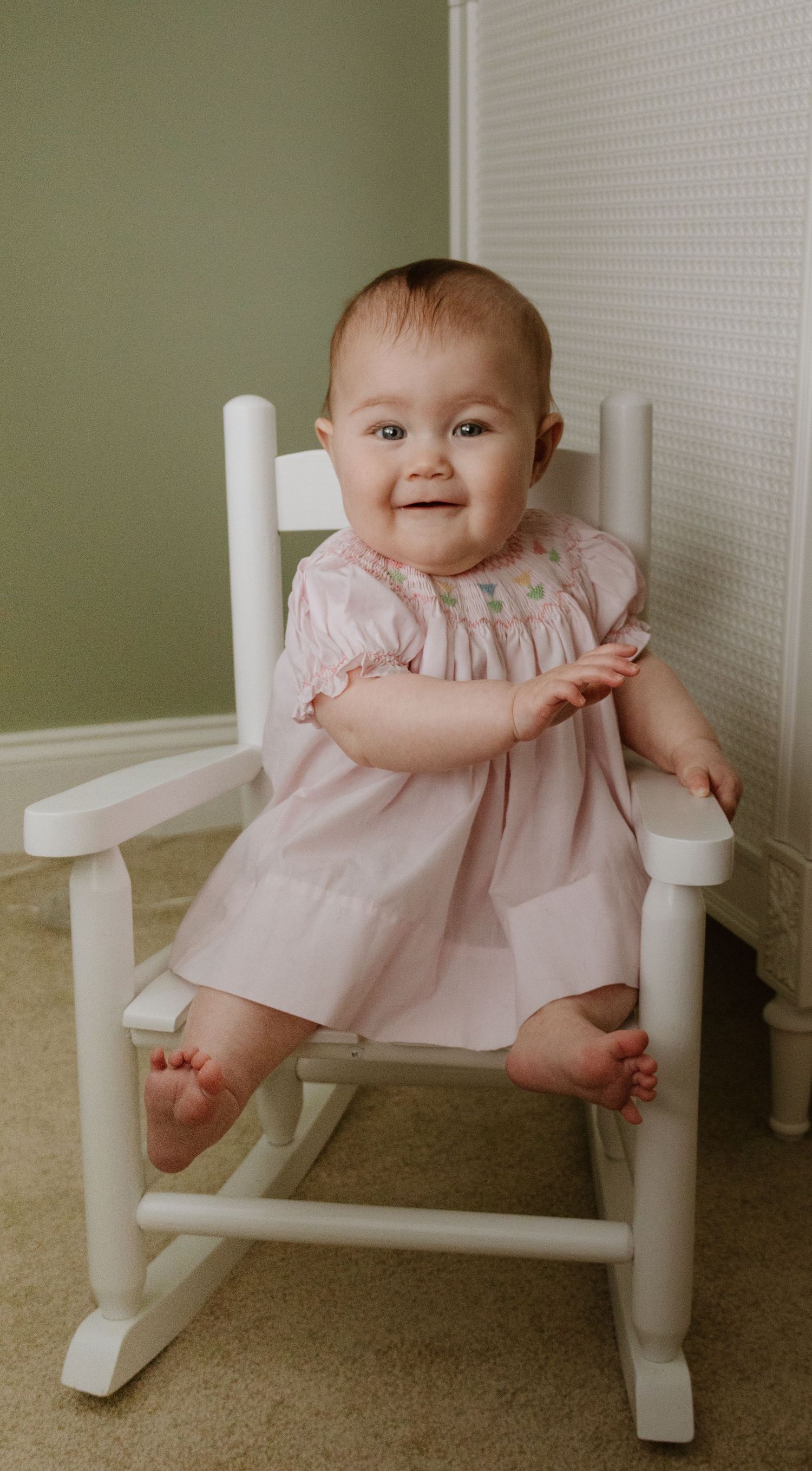 Baby in pink dress smiling while sitting in white wooden rocking chair.