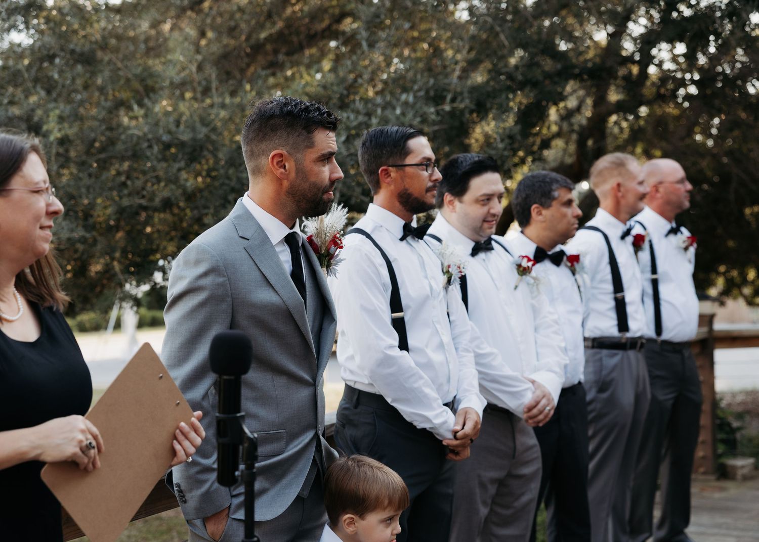 Groom in gray suit standing with groomsmen in white shirts and suspenders during outdoor wedding ceremony.