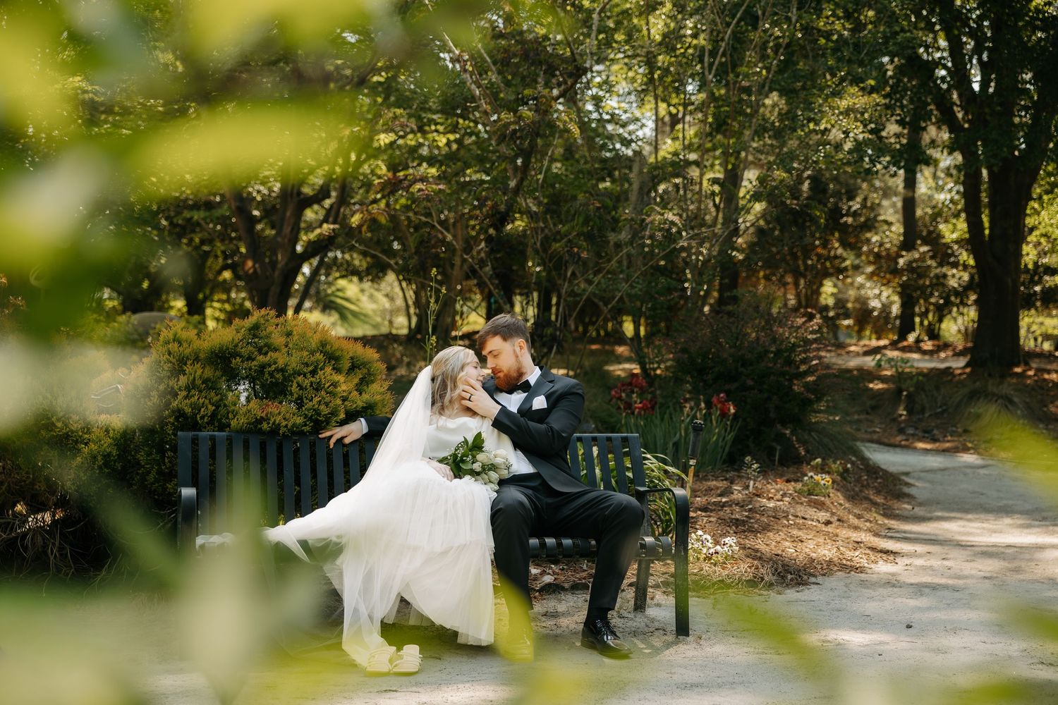 Bride and groom share a romantic kiss on a park bench surrounded by lush green trees on their wedding day.