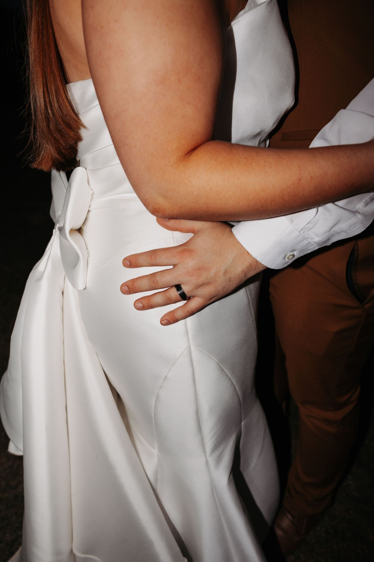 Close-up of wedding couple embracing, bride in white satin gown with bow, groom wearing black wedding band.