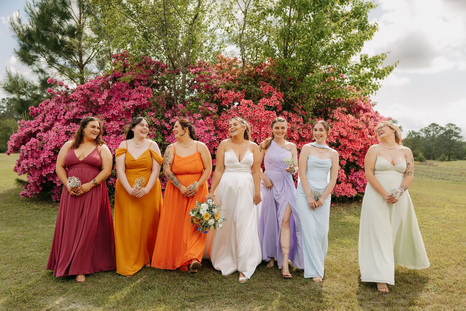 Bride and bridesmaids in rainbow-colored gowns pose outdoors in front of vibrant pink azalea bushes.