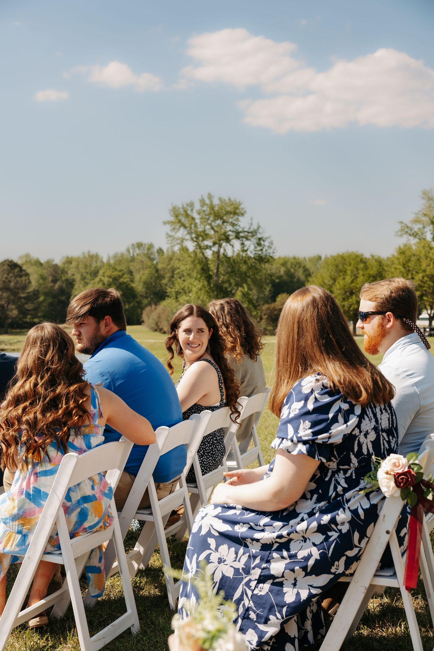 Guests seated in white chairs at an outdoor wedding ceremony on a sunny day with green trees in the background.