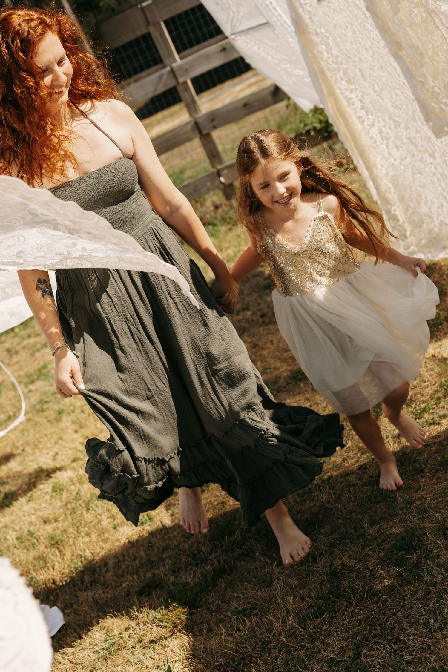 A redheaded woman in a green dress holds hands with a smiling girl in a tulle dress, dancing barefoot outdoors.