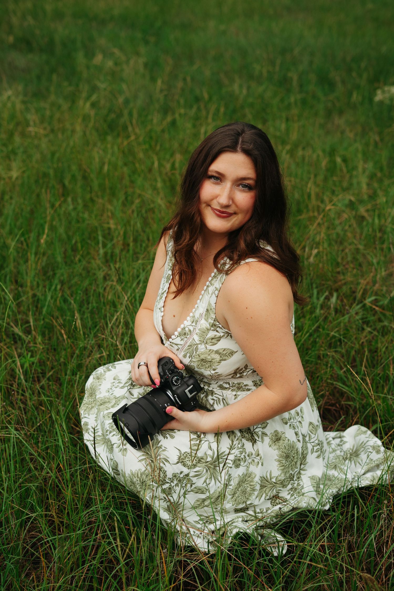 Woman in floral dress sitting in green field holding a camera, smiling outdoors.