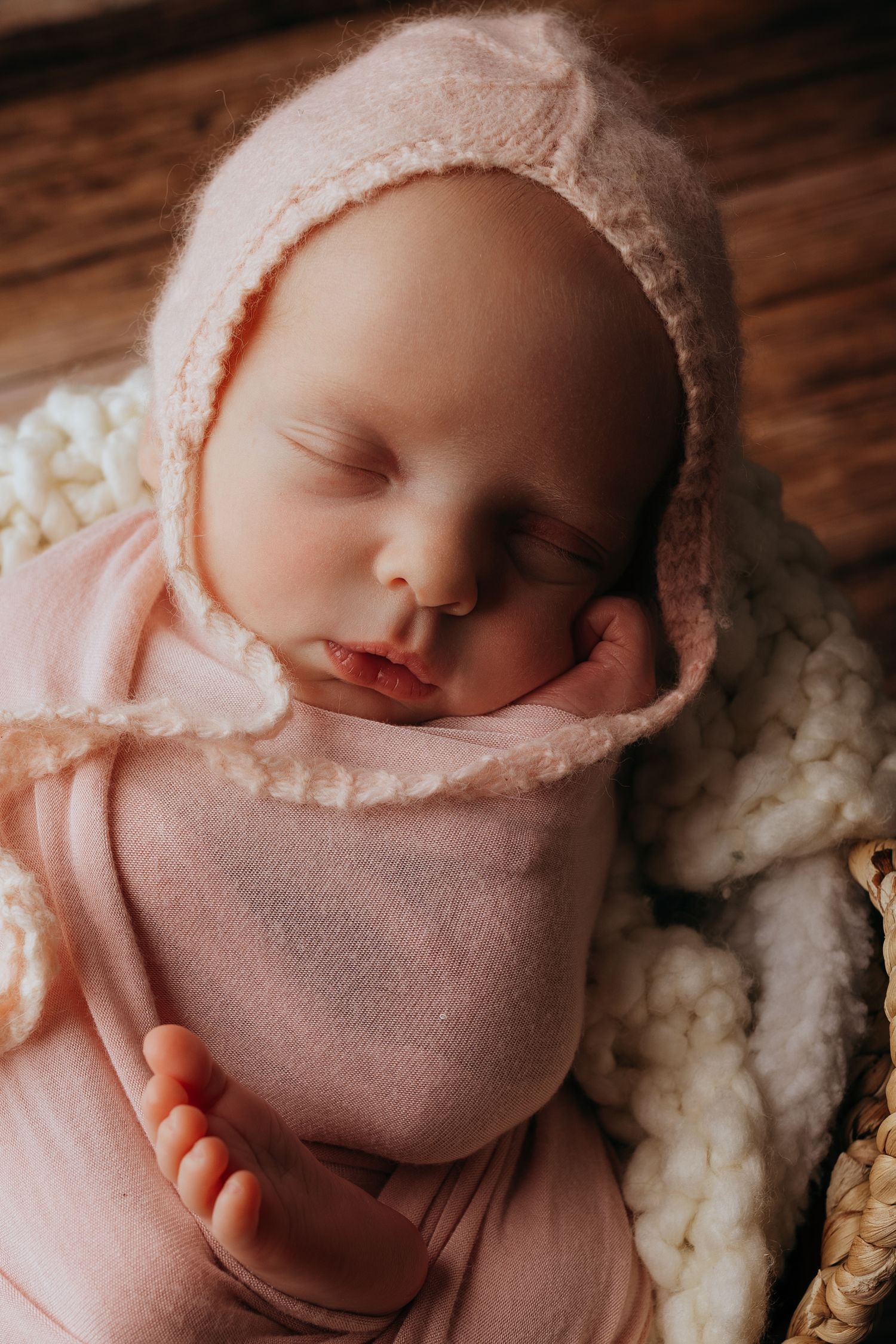 A sleeping newborn swaddled in soft pink fabric rests peacefully against a cozy white textured backdrop.