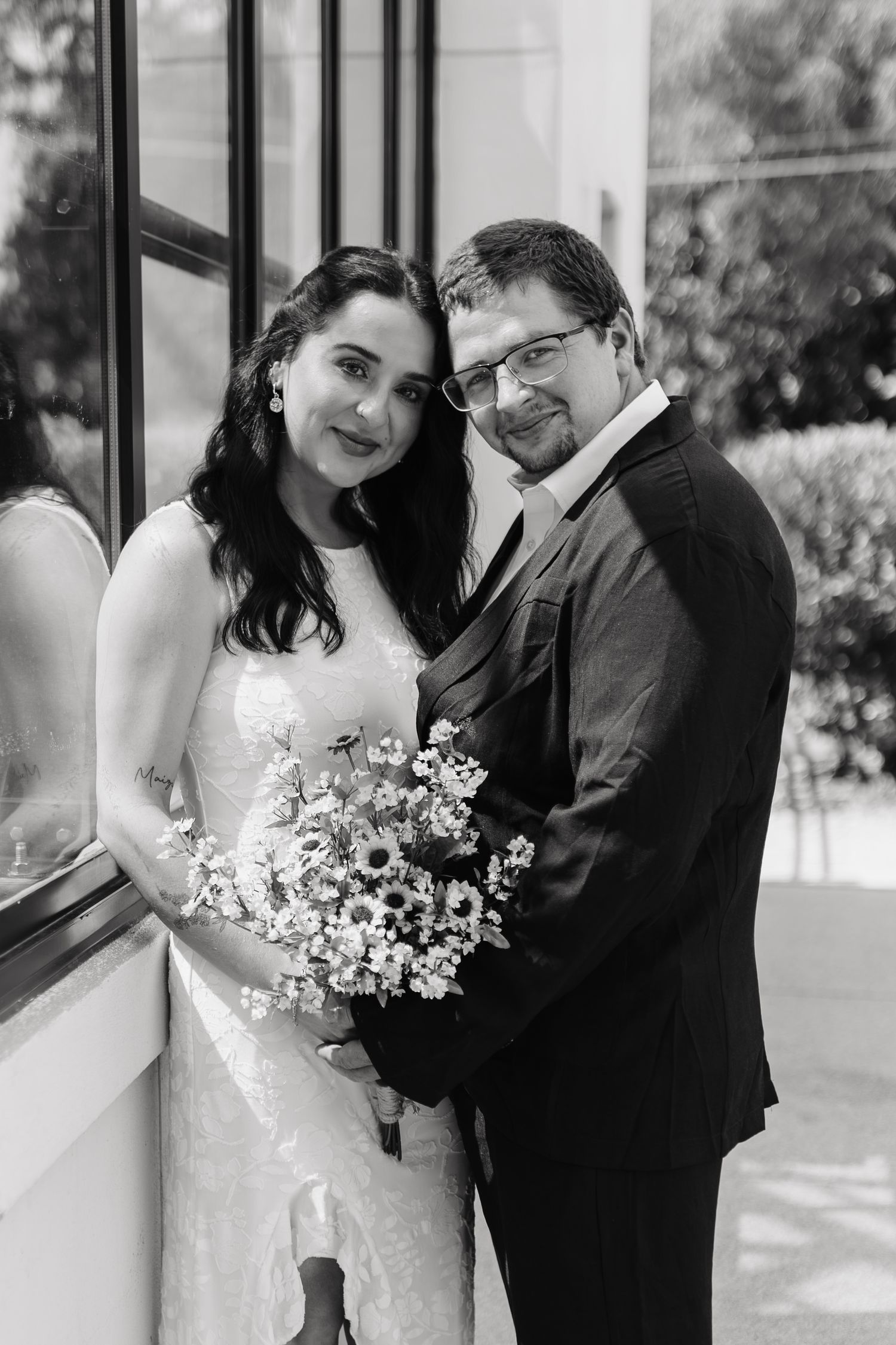 Black and white wedding portrait of bride holding floral bouquet and groom in suit posing together near window.