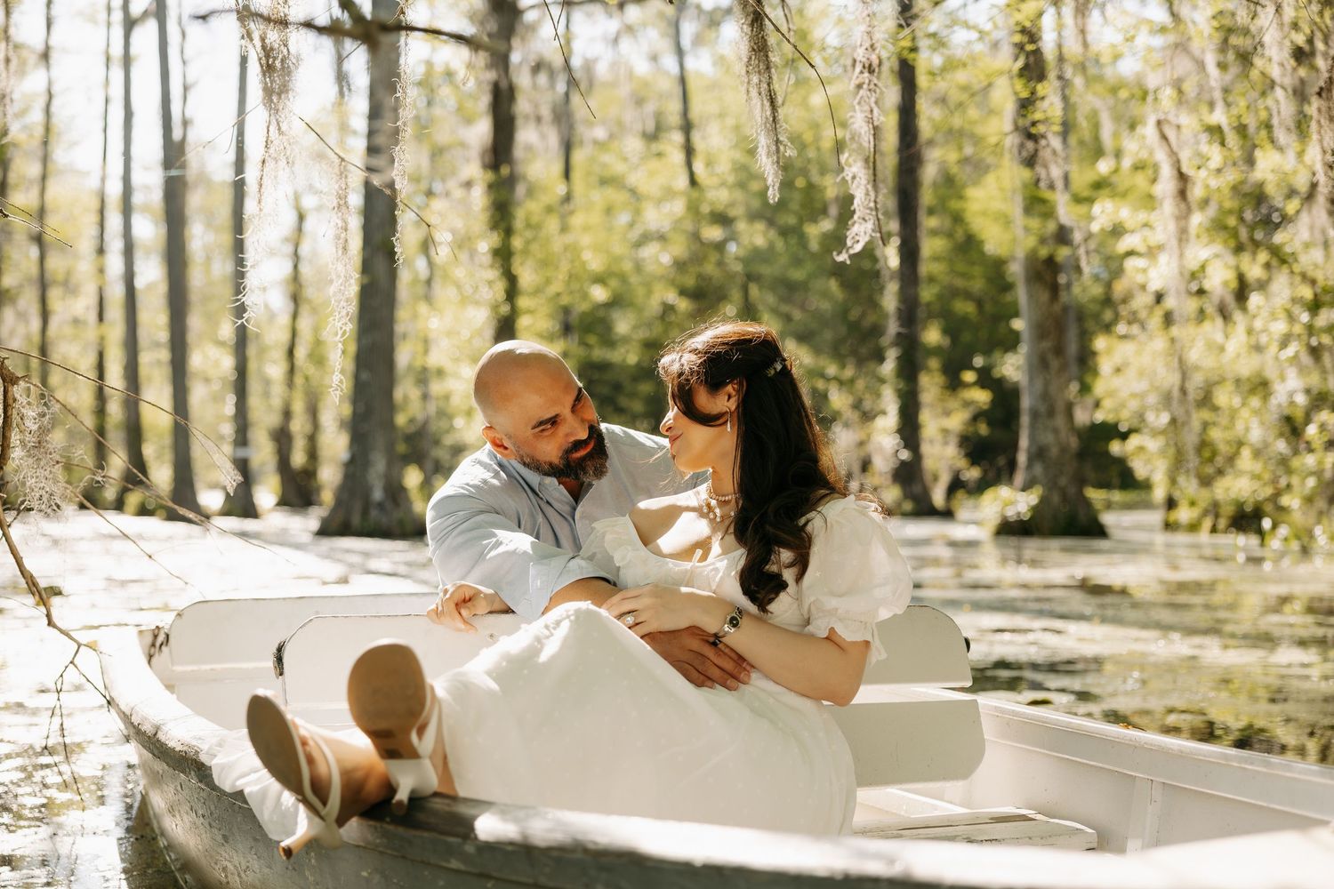 Couple sitting together in a white boat surrounded by lush green trees, sharing a romantic moment.