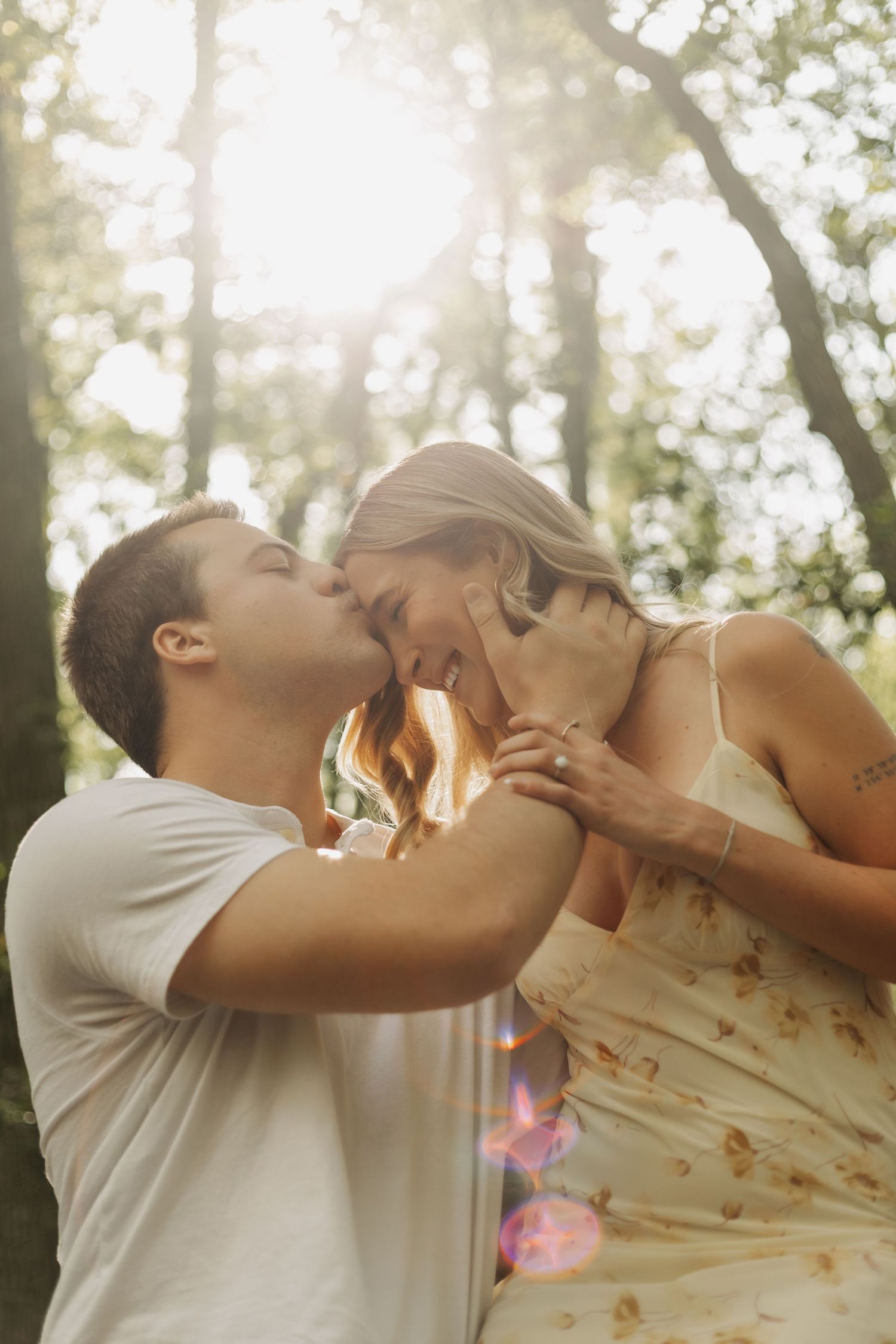 A couple shares a romantic kiss outdoors in sunlit forest, woman in floral dress holding mans face tenderly.