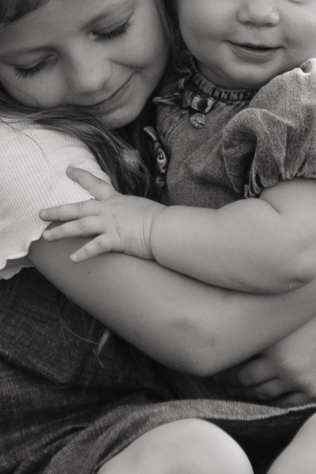 Black and white photo of an older girl lovingly hugging a smiling baby sibling.