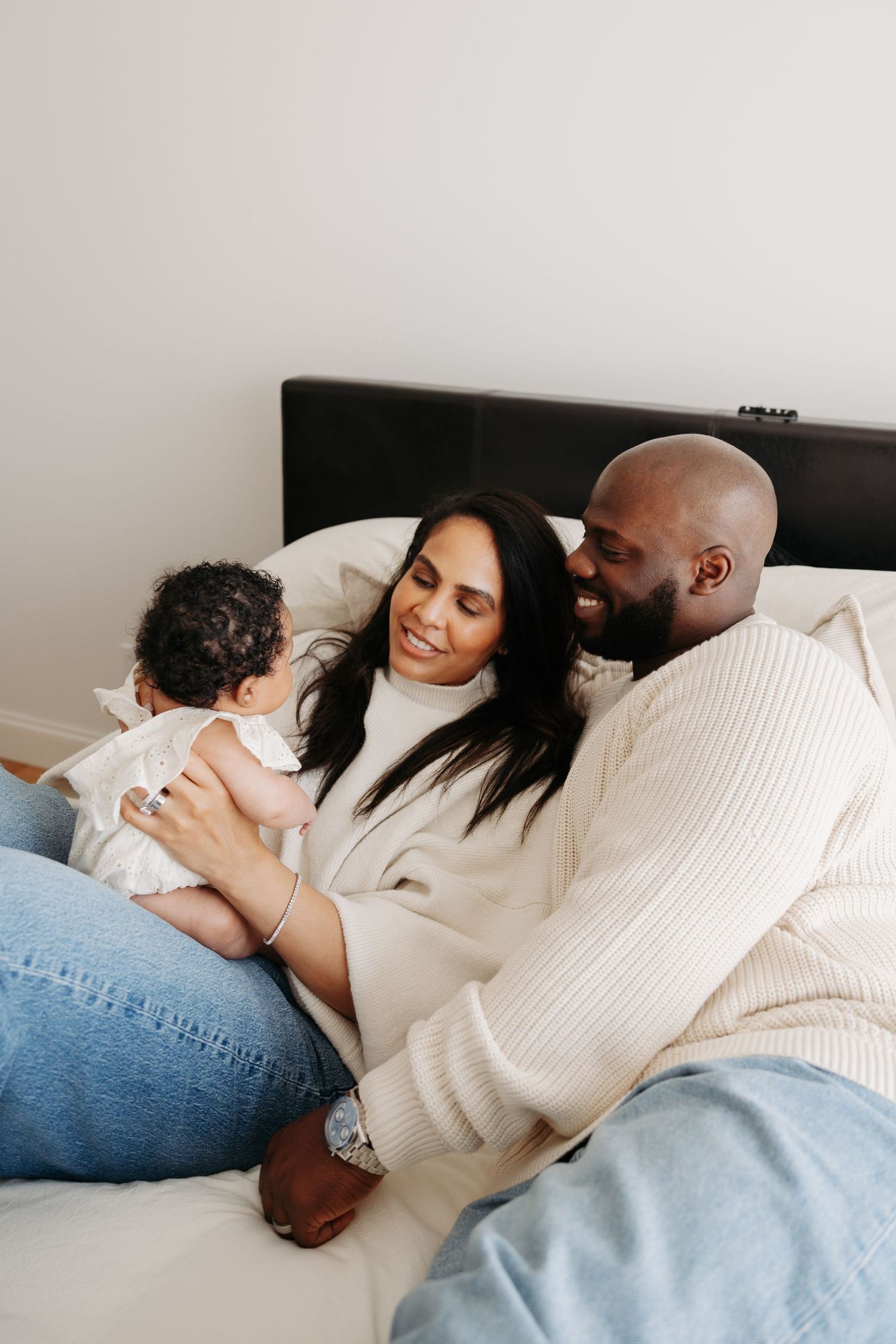 A happy couple sits on a bed smiling at their newborn baby, sharing a warm family moment together.