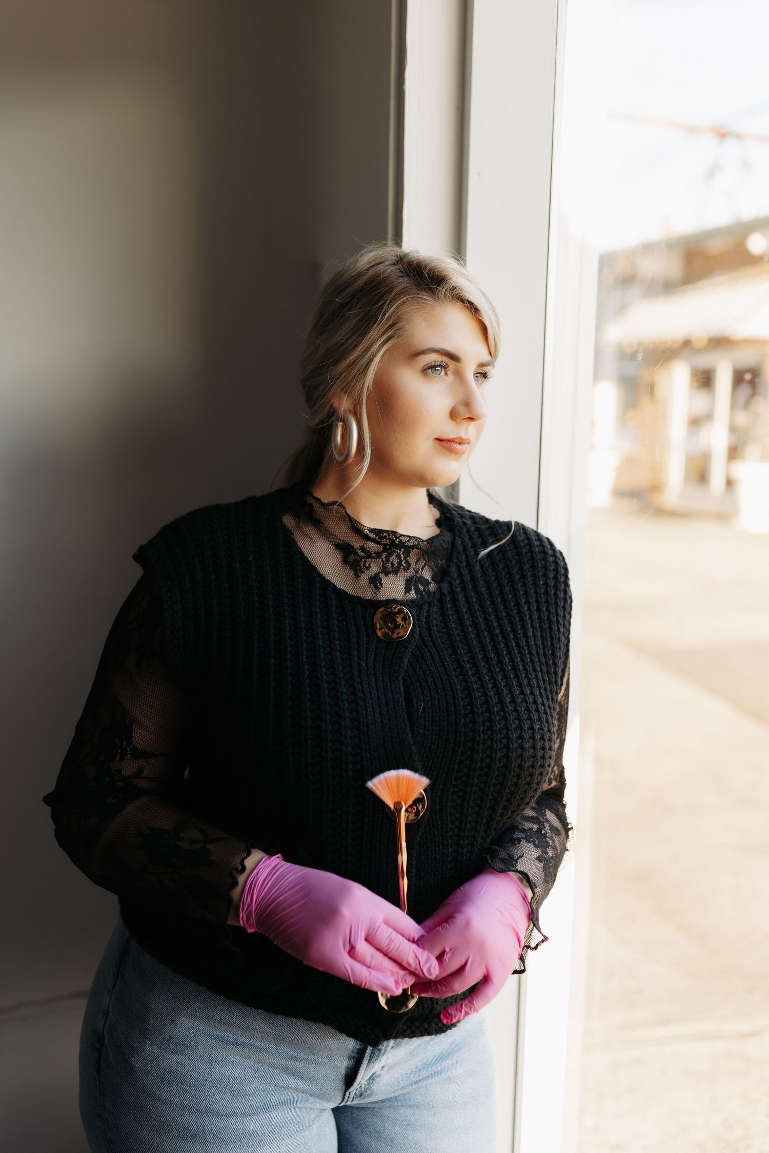 Person wearing black sweater and pink gloves poses by window in natural lighting.