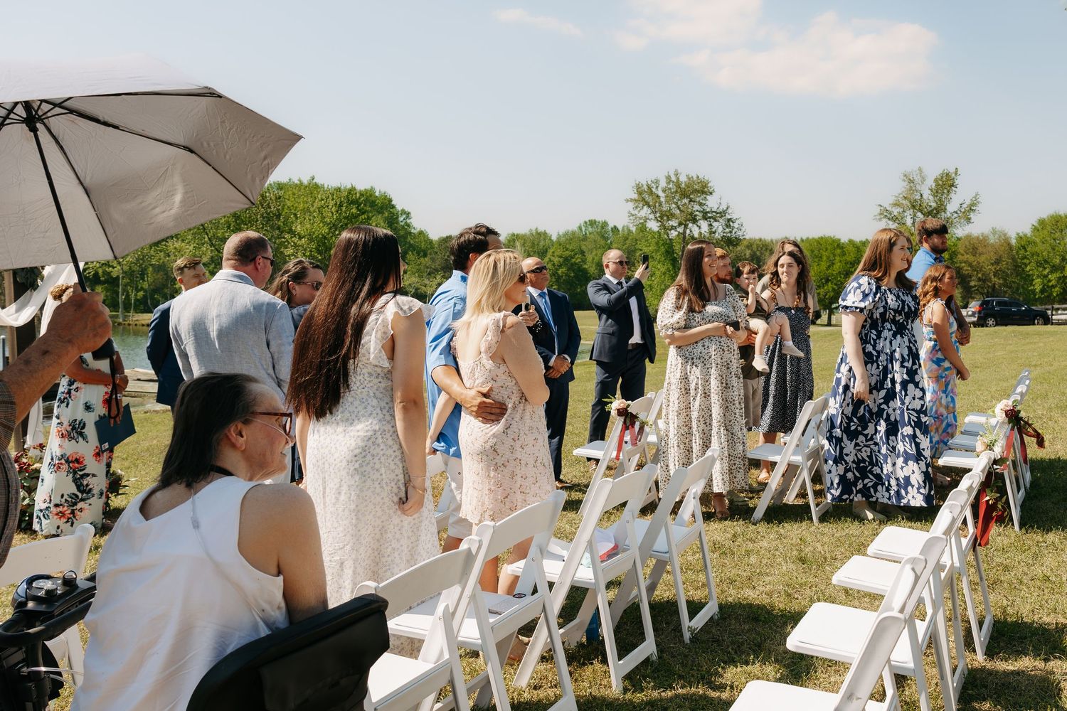 Guests mingling outdoors at a sunny wedding ceremony with white chairs on green grass.