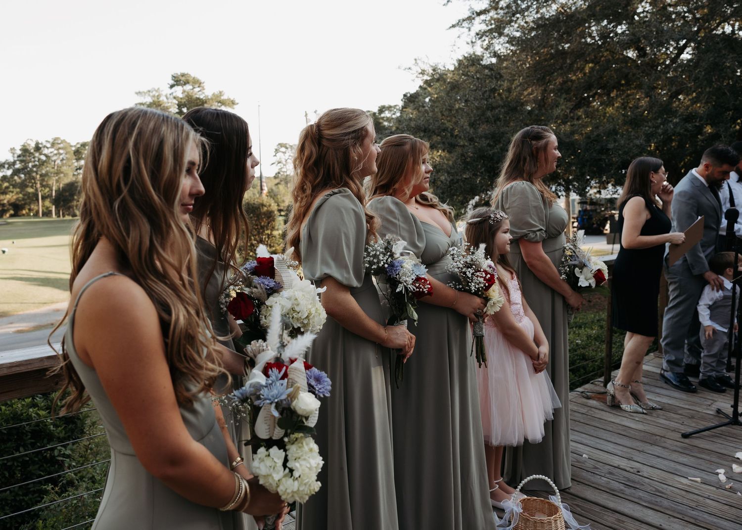 Bridesmaids in sage green dresses holding bouquets standing at an outdoor wedding ceremony.