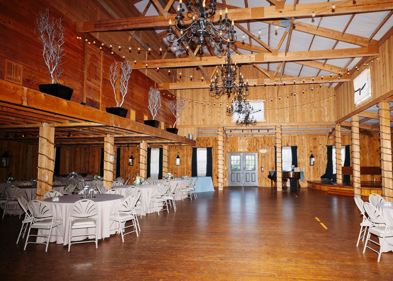 Rustic wedding reception hall with wood walls, chandeliers, white-draped tables, and string lights in a barn venue.