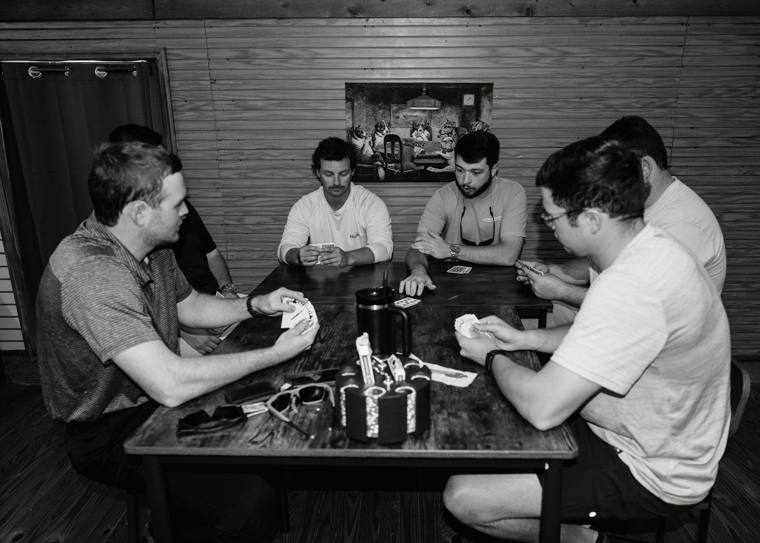 Five men playing cards at a wooden table in a rustic room, black and white photo.