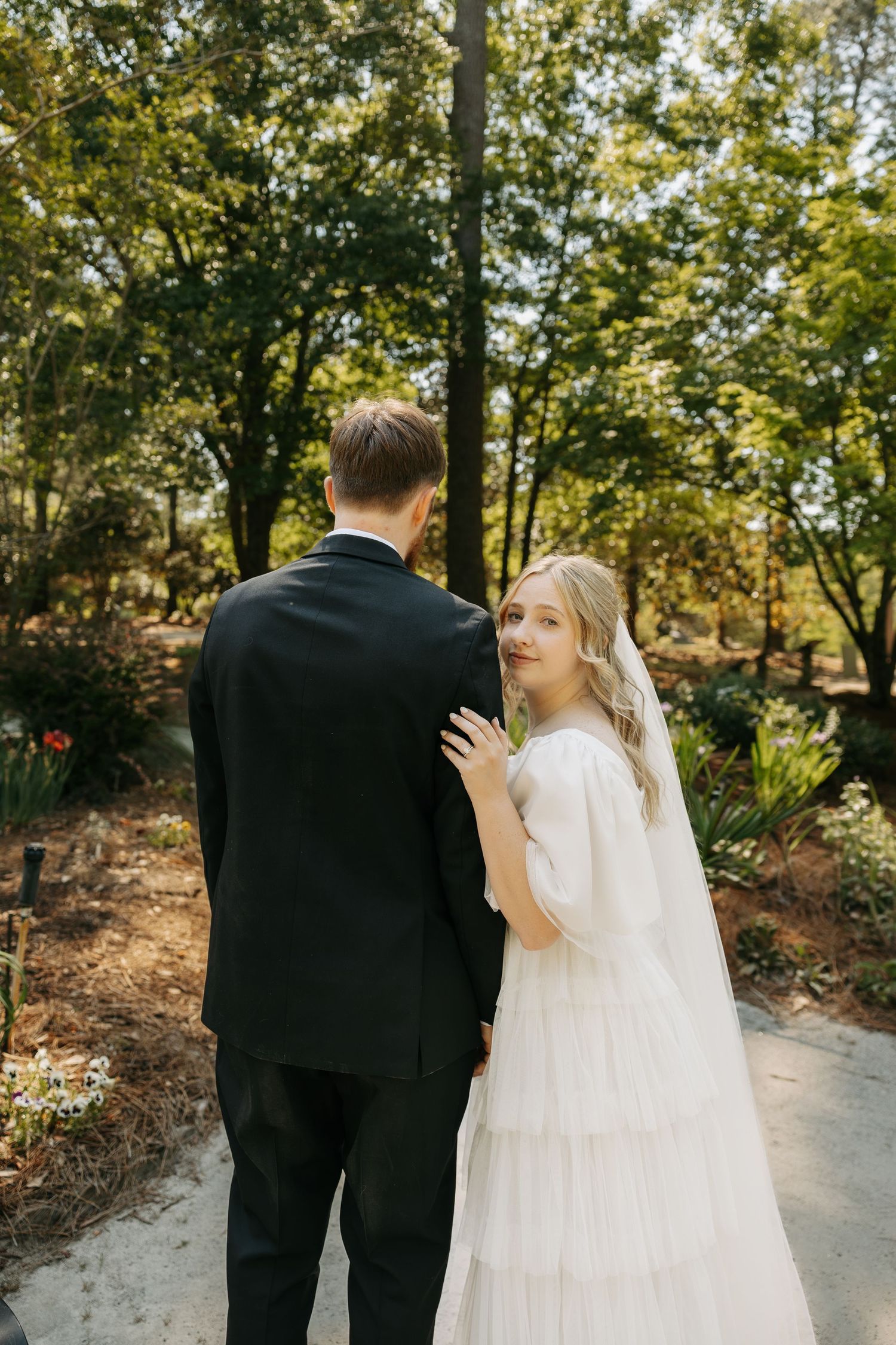 Bride in white tiered dress glancing back while walking with groom in garden.