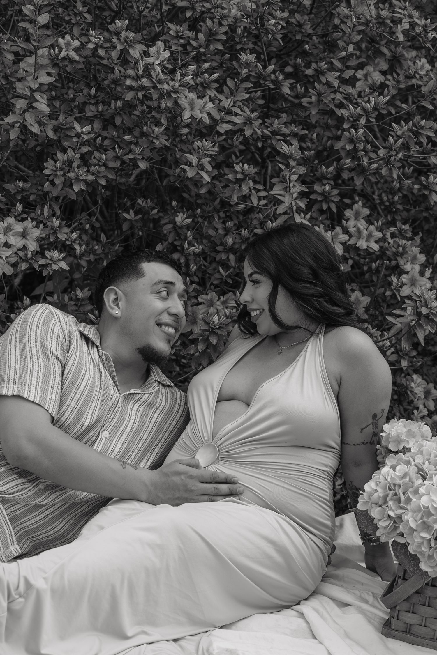 Black and white photo of a smiling couple sitting together outdoors near flowering bushes, woman in maternity dress.