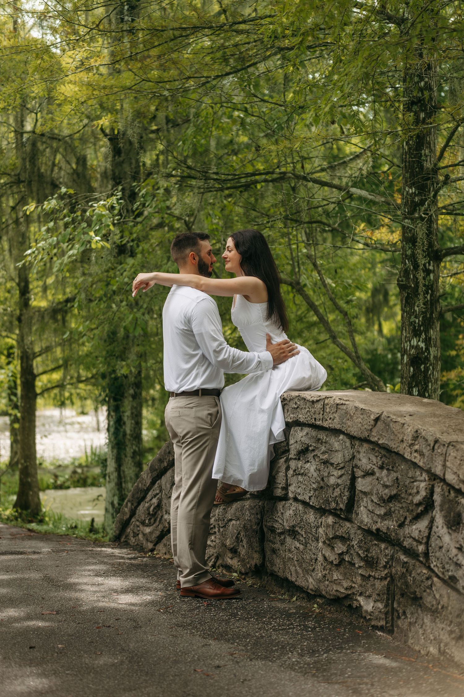 Romantic couple embracing on a stone bridge in a lush green forest, woman in white dress seated on wall.