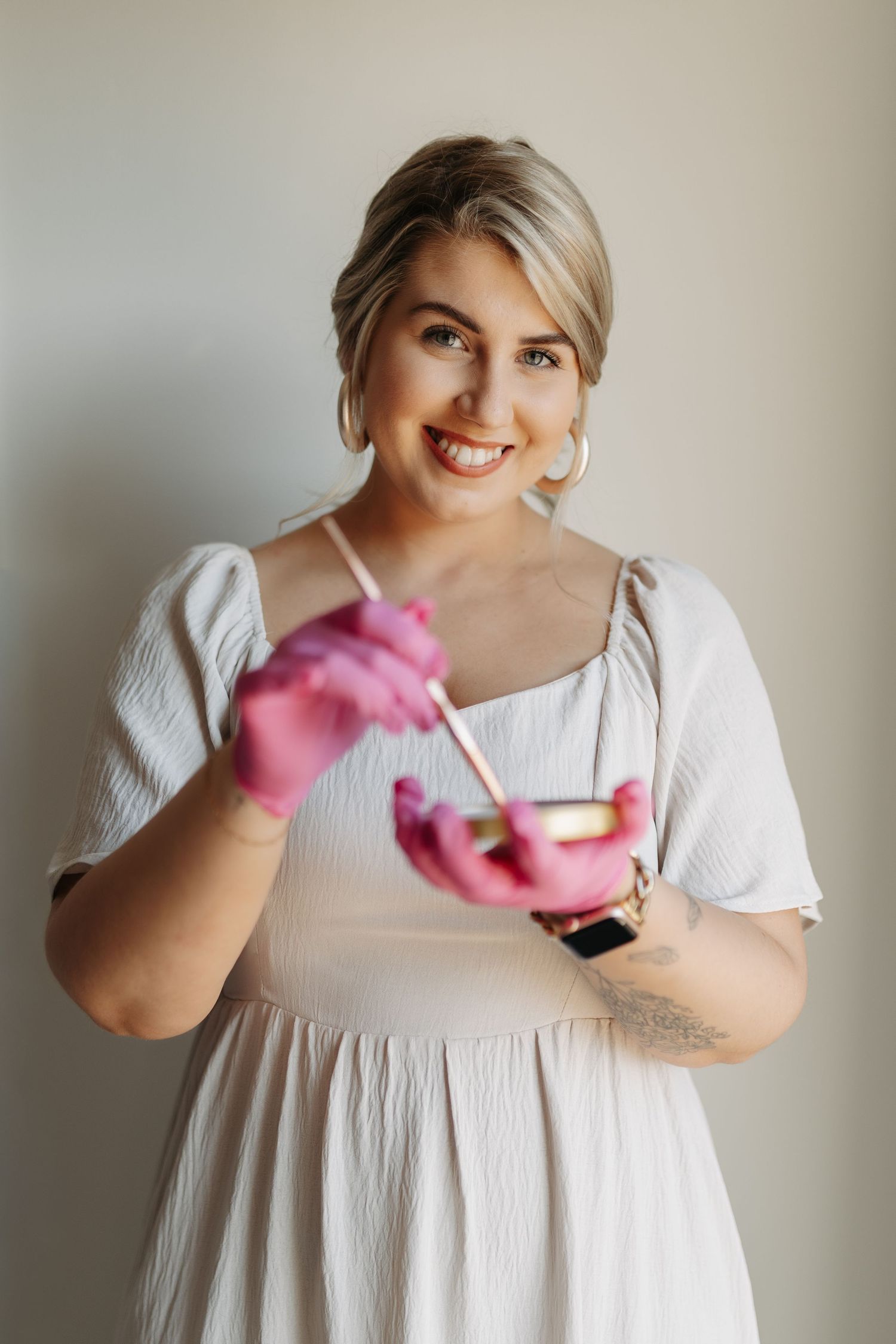Person in white dress putting on pink cleaning gloves while standing against neutral background.