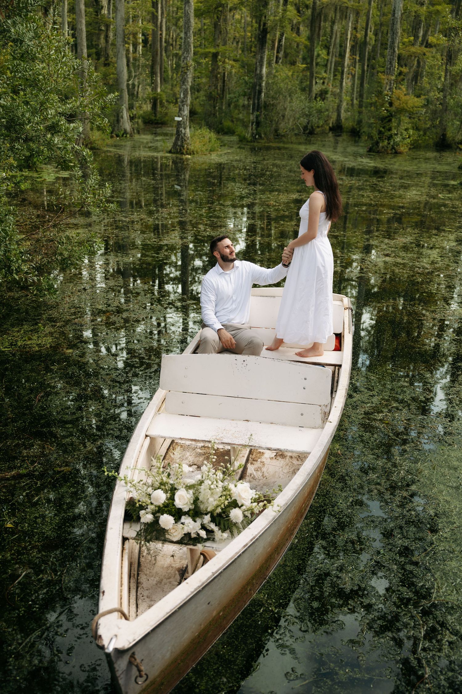 Couple in white attire sharing a romantic moment on a rustic wooden canoe surrounded by a lush green forest lake.