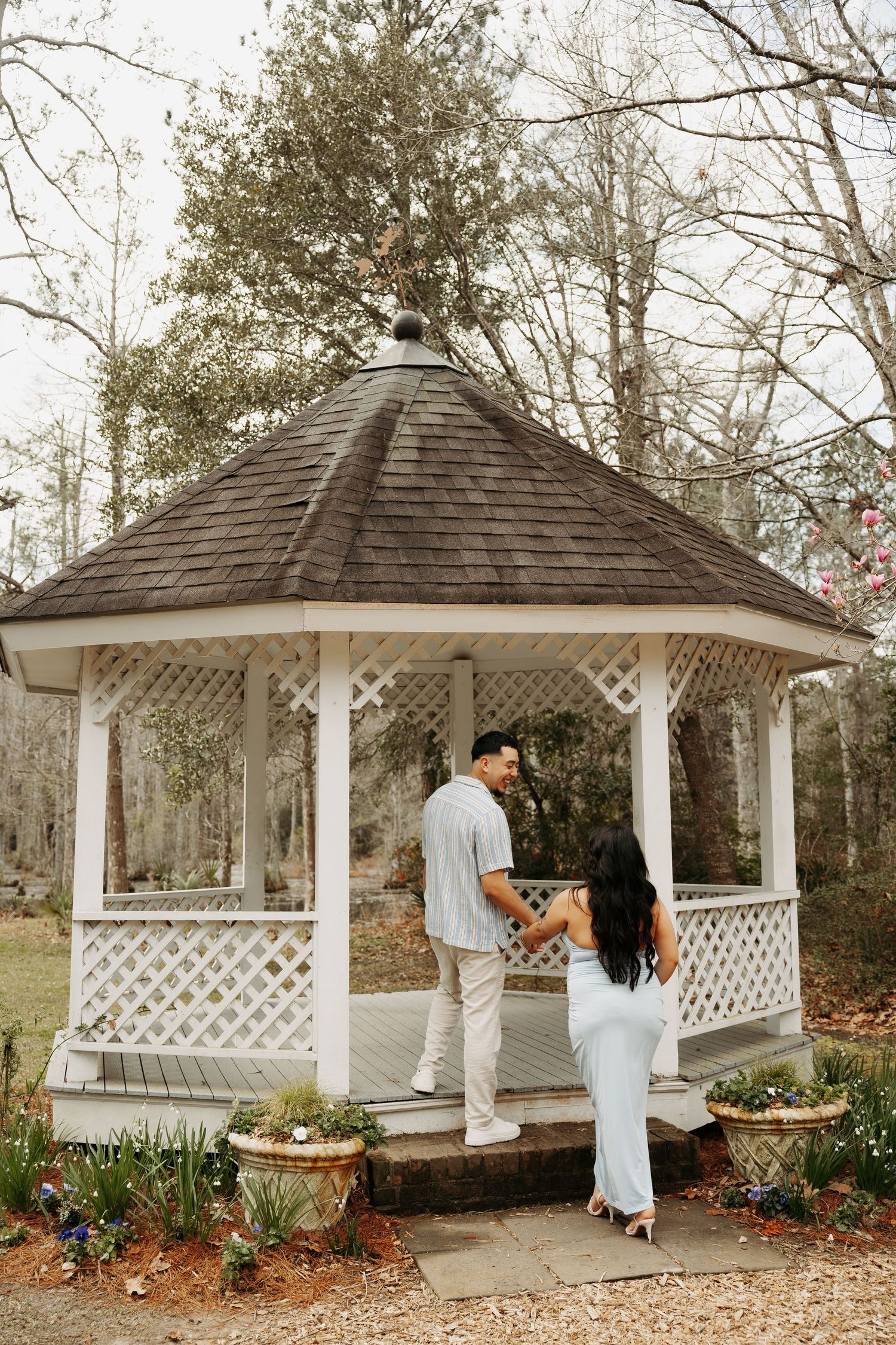 Couple holding hands at a white wooden gazebo surrounded by trees in a garden setting.