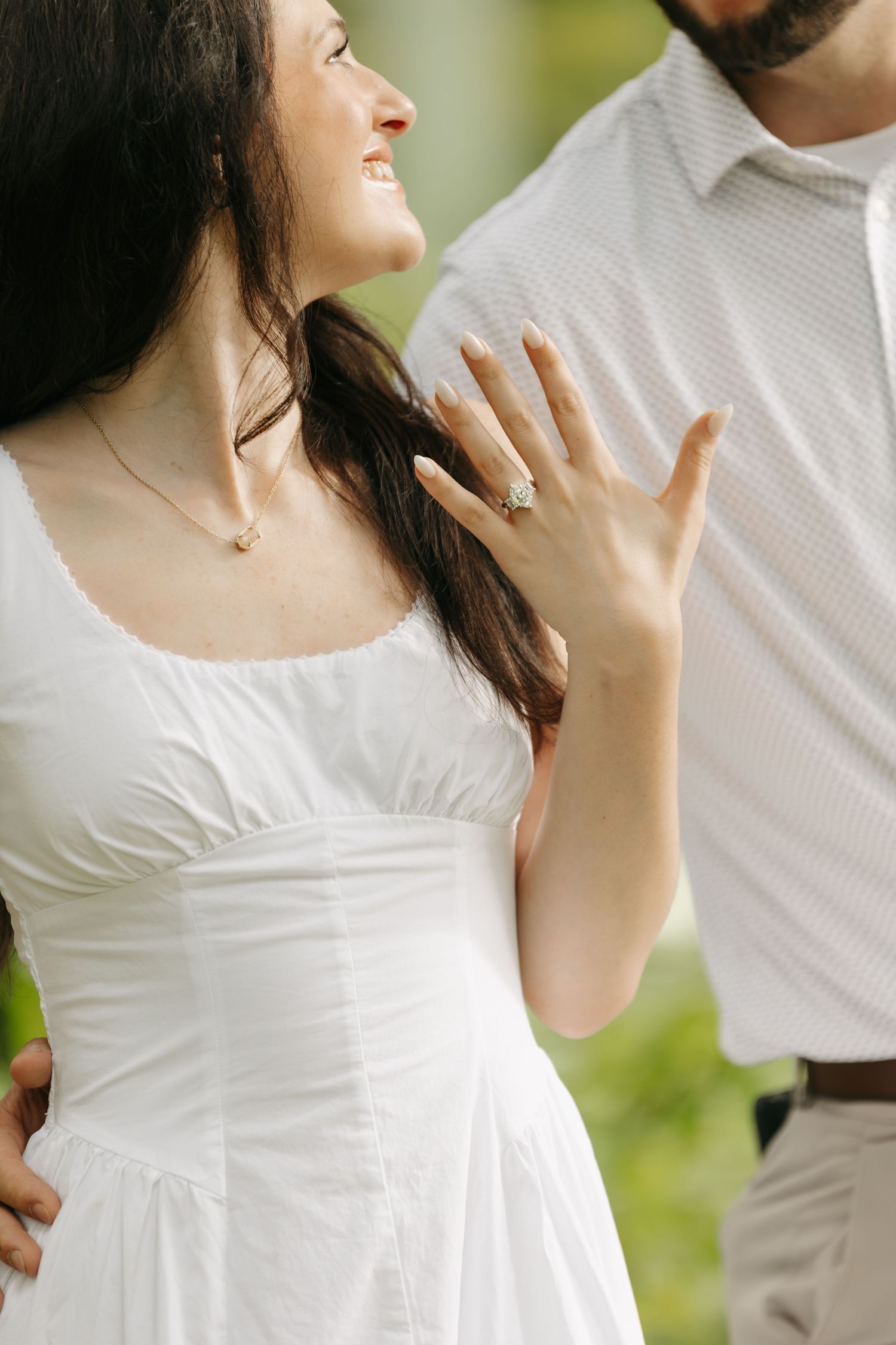 Smiling bride in white dress proudly displays engagement ring while standing with groom outdoors.