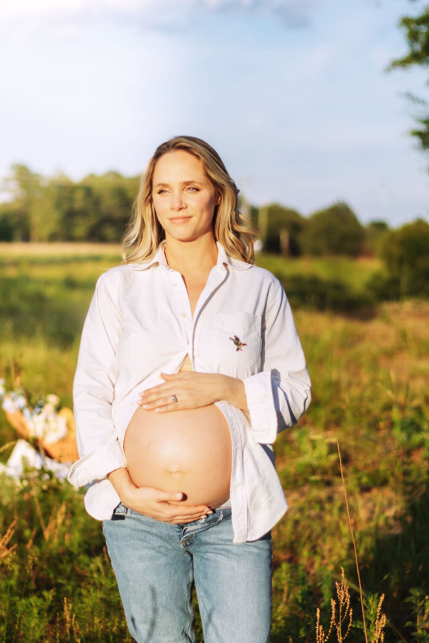 Pregnant woman in white shirt and jeans cradling baby bump in sunny field.