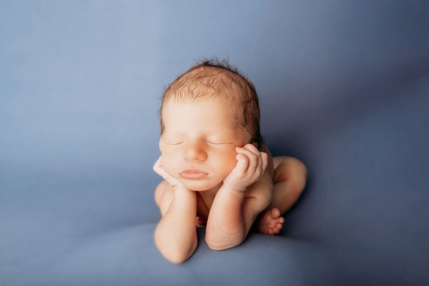 Peaceful newborn baby sleeping curled up on soft blue fabric, hands tucked under chin in classic posed newborn photography.