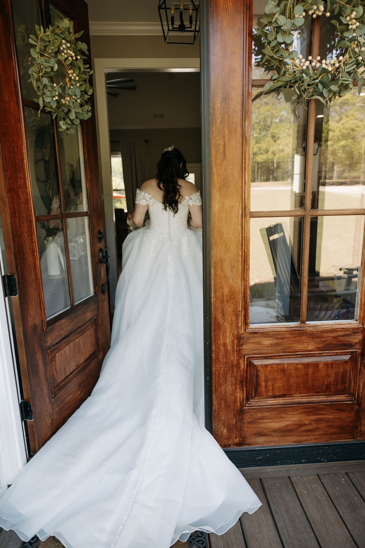 Bride in white off-shoulder gown with long train stands at wooden double doors adorned with greenery wreaths.