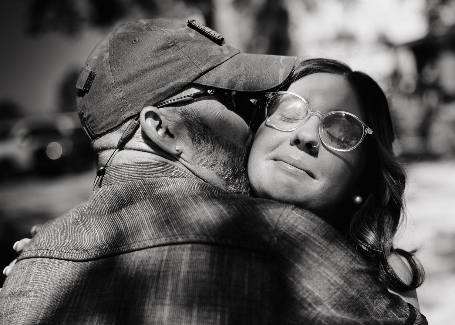 Black and white photo of a woman with glasses hugging a man in a plaid shirt and cap outdoors.