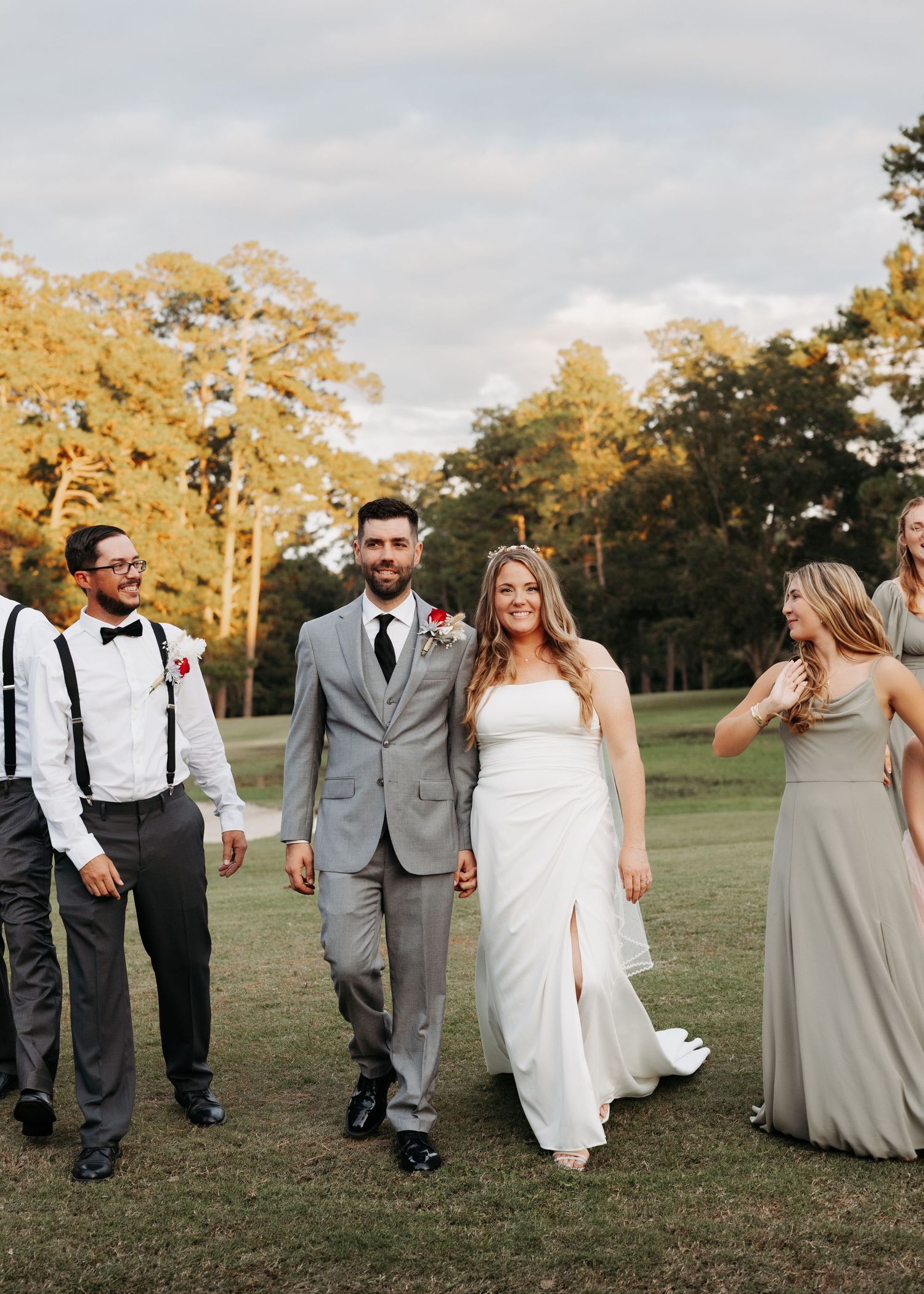 Bride and groom walk hand in hand with wedding party on grassy field surrounded by autumn trees at outdoor wedding.